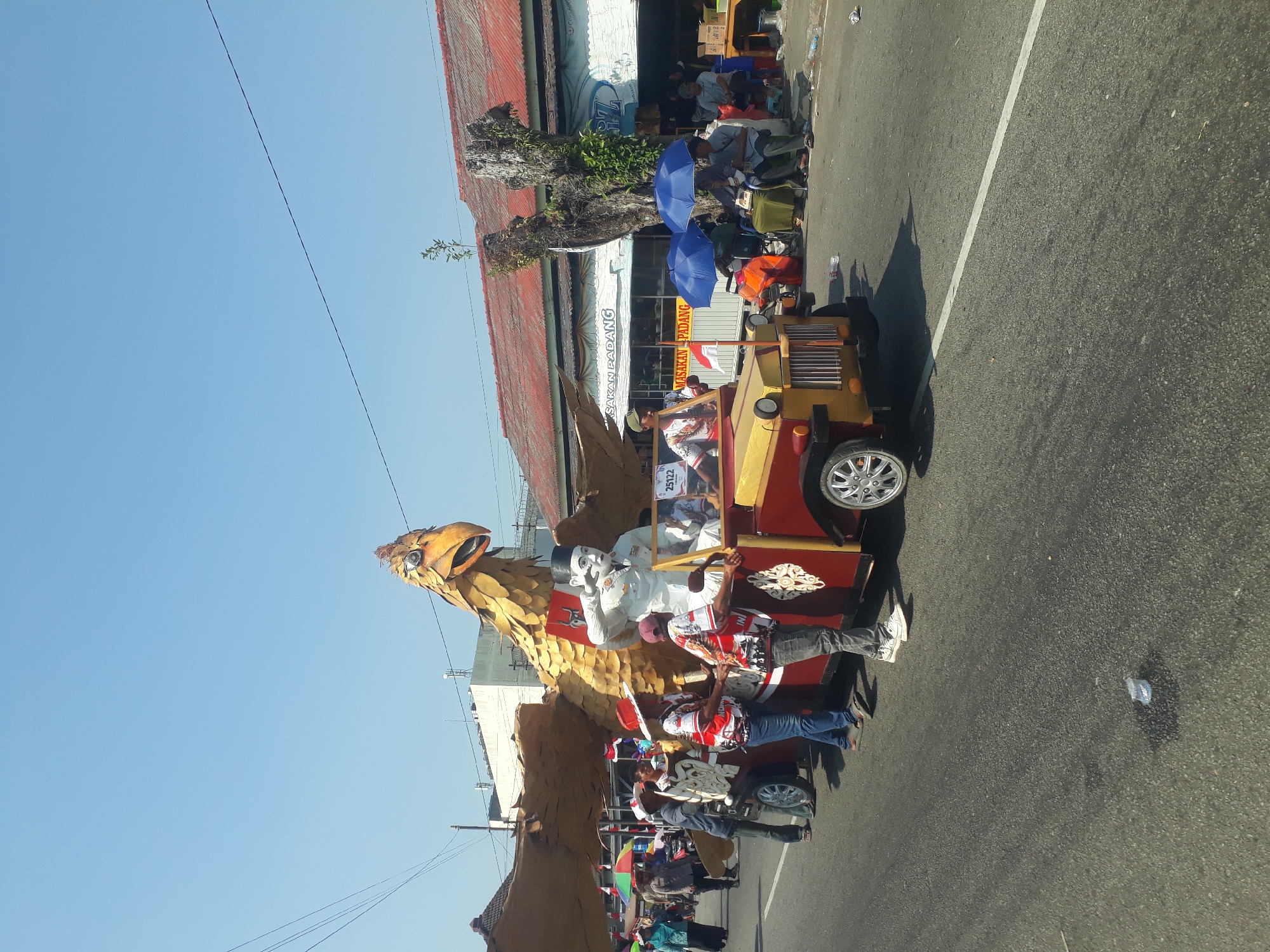 A parade float, designed as a golden eagle with outstretched wings and a human effigy in military attire saluting, is being pulled by a red and yellow jeep-like vehicle along a street in Pangkalpinang, Indonesia. Several individuals, some wearing matching red and white patterned shirts, are accompanying the float, pushing or guiding it. Spectators are visible on the sidewalks, some sitting under blue umbrellas. Shops with awnings line the street. The sky is clear and blue, indicating a sunny day.