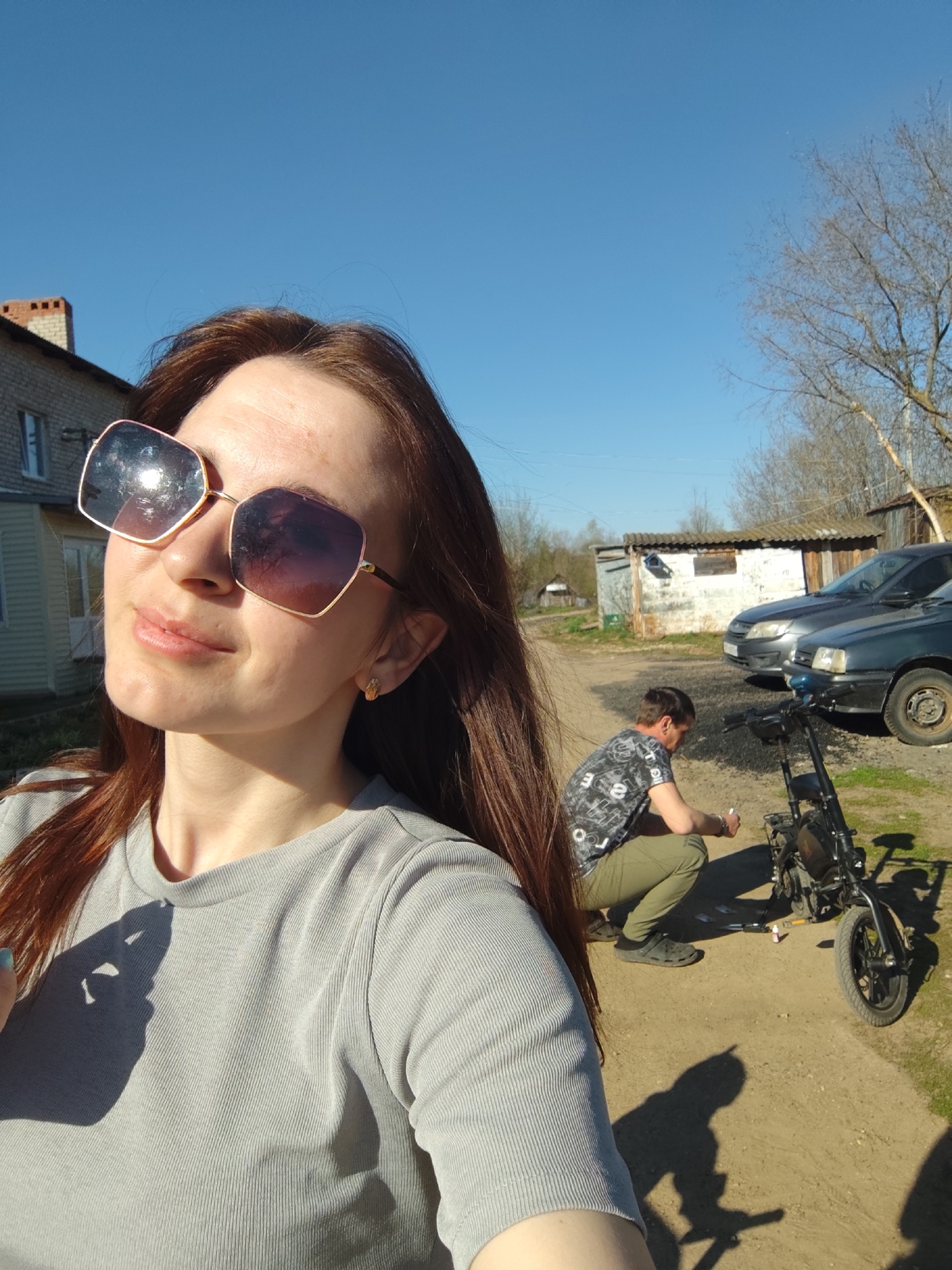 Outside, a woman with long, brown hair, wearing a gray t-shirt and large, rose-gold sunglasses, is taking a selfie. In the background, a man is squatting next to a small, black motorized bicycle, appearing to work on it. Two cars are parked behind him, and there are a few sheds and a house visible. The location is Фоки, Russia, as the model is instructed. The sky is clear and blue. The ground is dirt and gravel.