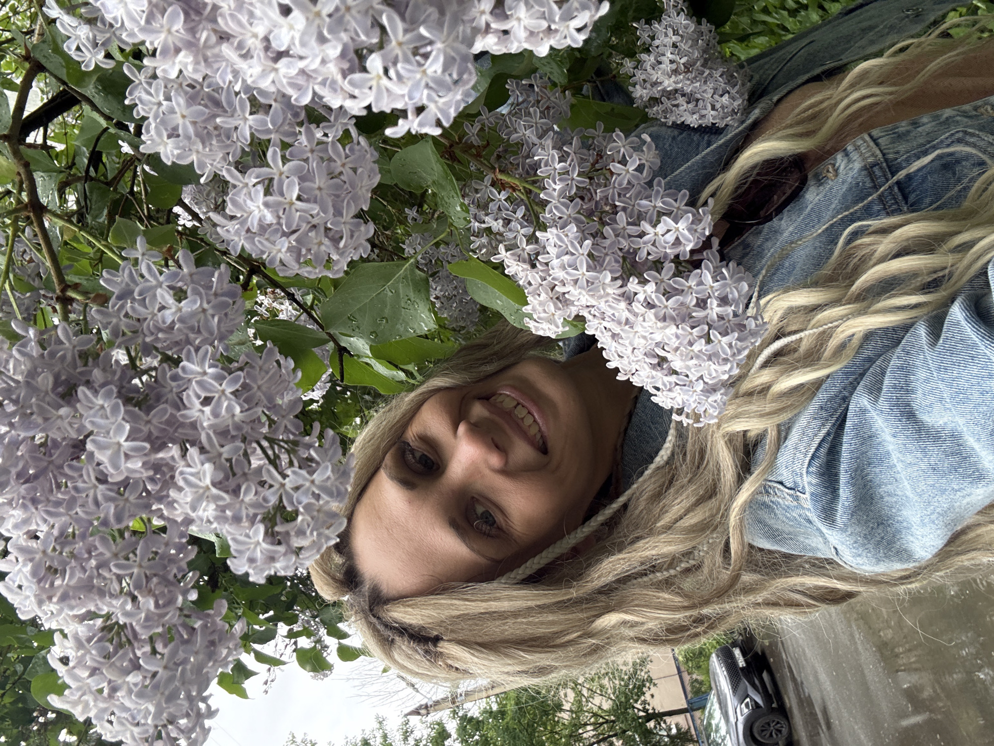 A woman with blond hair smiles while surrounded by lilac blossoms. She wears a denim jacket. A vehicle is visible in the lower right corner, parked on a wet surface suggesting it may have rained. Based on the available information, it's not possible to confirm the exact location or interactions specifically in Rusanovo, Russia. However, the scene appears to be outdoors, near flowering trees with a vehicle parked nearby, suggesting an urban or suburban environment.
