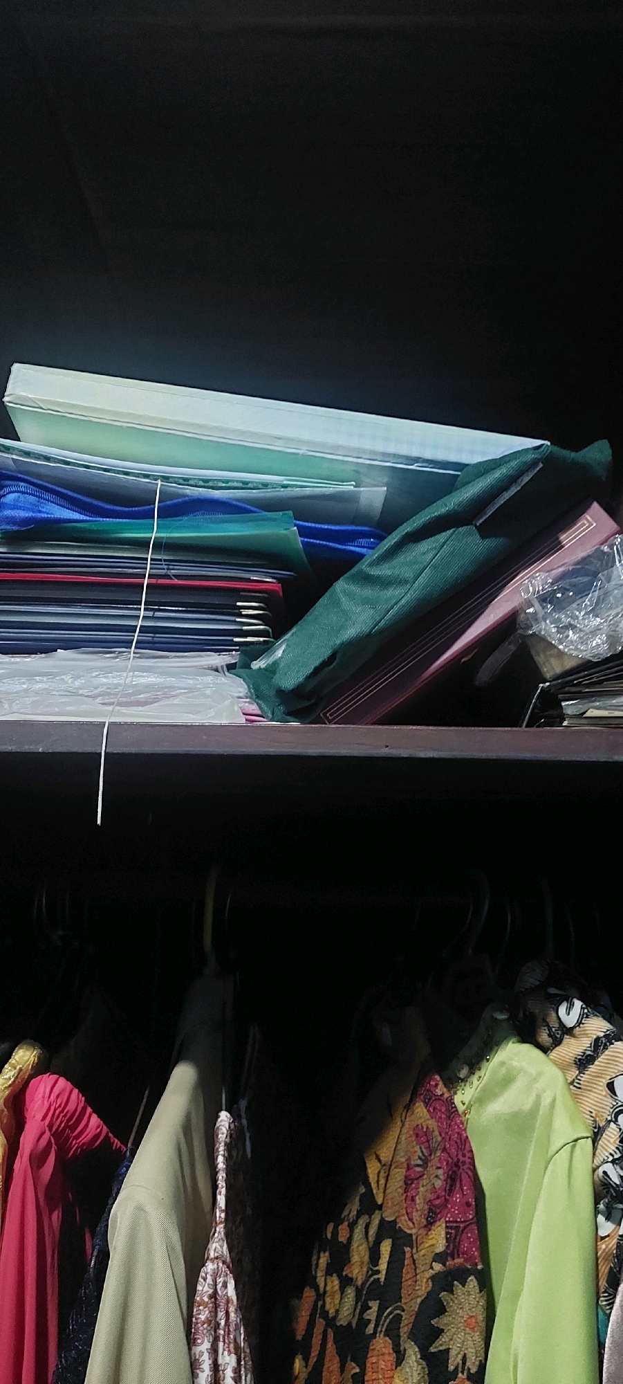 The image shows the inside of a dark wardrobe with two shelves. The top shelf is filled with files and folders, some of which are green and blue. A white string dangles from the files. On the lower shelf, clothes are hanging on hangers, including a pink dress, a patterned fabric, and a green shirt. The wardrobe is located in Lumajang, Indonesia.