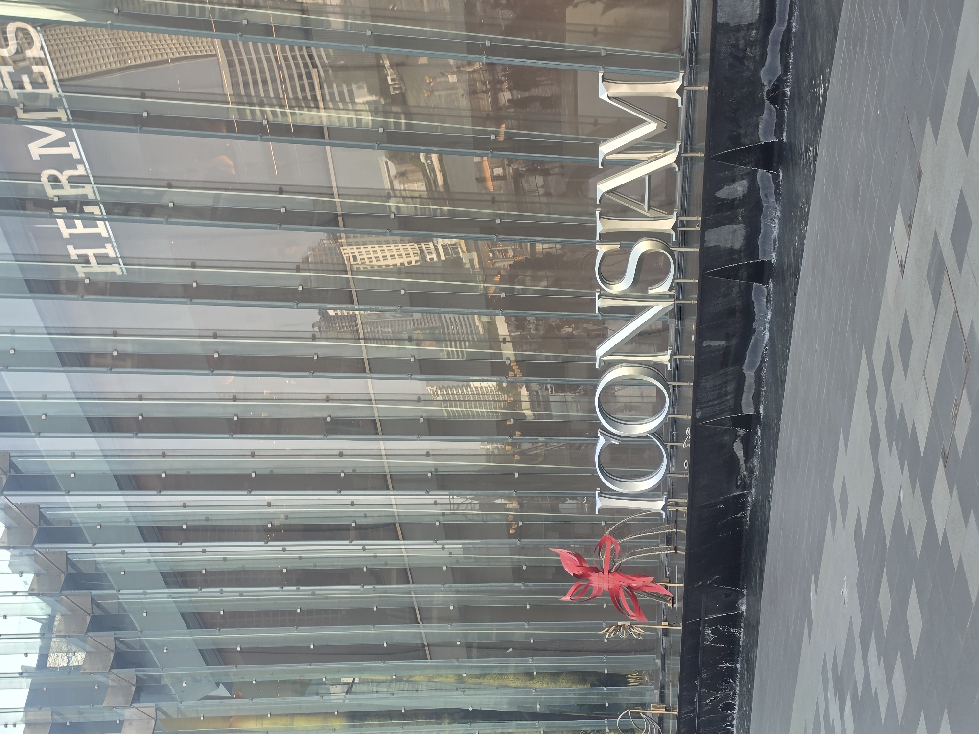 The image shows the ICONSIAM shopping mall in Bangkok, Thailand. The facade of the building has the "HERMES" brand and "ICONSIAM" written on it. The building has a unique architectural style. There is a red decoration in front of the ICONSIAM sign. There is a water feature along the side of the building that looks like a man-made waterfall. The surrounding area has a gray concrete-tiled surface. The reflections of surrounding buildings are visible on the glass facade of ICONSIAM.