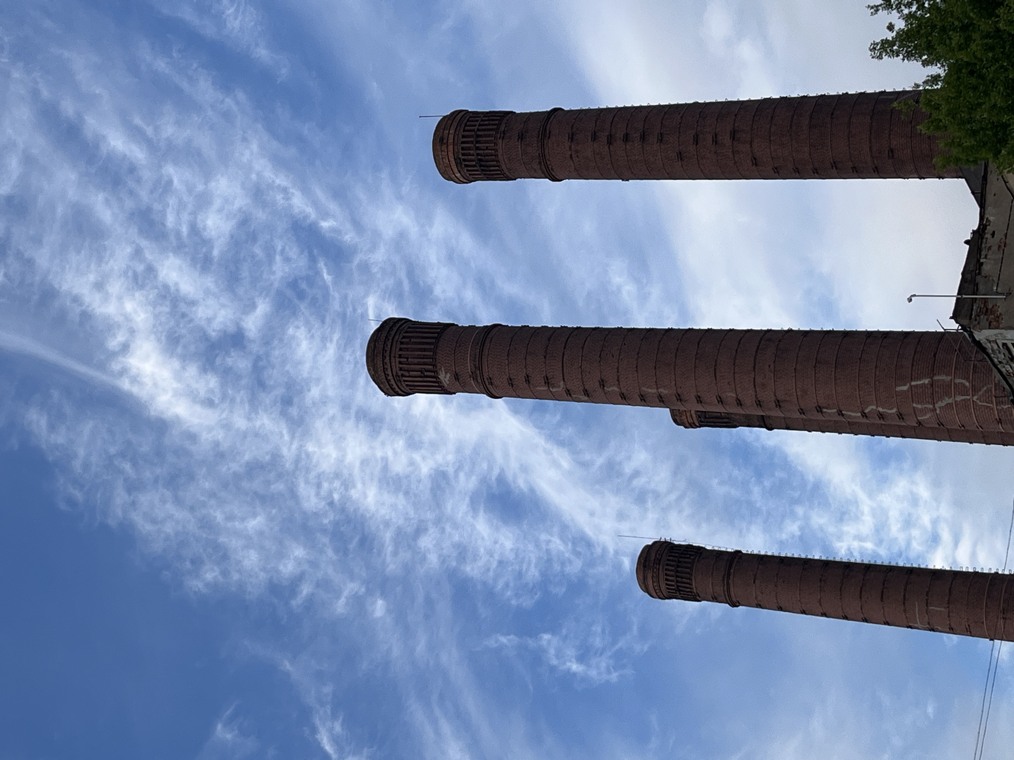 An upward-looking photograph taken in Saint Petersburg, Russia, captures three massive industrial chimneys set against a vibrant blue sky dotted with delicate, wispy white clouds. The cylindrical chimneys, constructed from reddish-brown brickwork, are positioned diagonally across the frame, stretching from the top right towards the bottom left, accentuating their immense scale. Each chimney culminates in a distinct, ribbed metal cap or collar, from which thin, dark wires—likely lightning rods—extend.
On the far right side of the image, the upper section of a building's facade or roofline is partially visible, featuring some dark green tree foliage at the extreme top right corner. A small, dark, pole-mounted object, possibly a light or sensor, is also discernible on the building structure. The bright sky and clear illumination indicate the photo was taken during the daytime under fair weather conditions. The scene is devoid of people or specific activities, presenting a static architectural landscape. There is no visible text in the image.