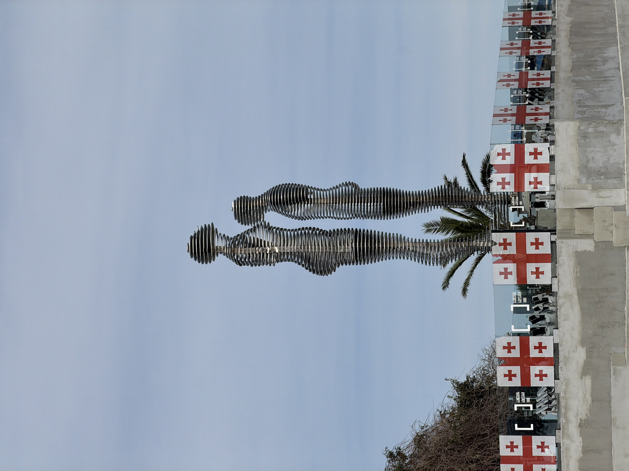 The image depicts the "Ali and Nino" statue in Batumi, Georgia. The statue consists of two abstract, steel-ringed figures representing a man and a woman. These figures are positioned under a clear, pale sky. The base of the statue is surrounded by vegetation. To the right, a building is adorned with Georgian flags. The photograph is taken from an unconventional angle, adding a unique perspective to the subject.