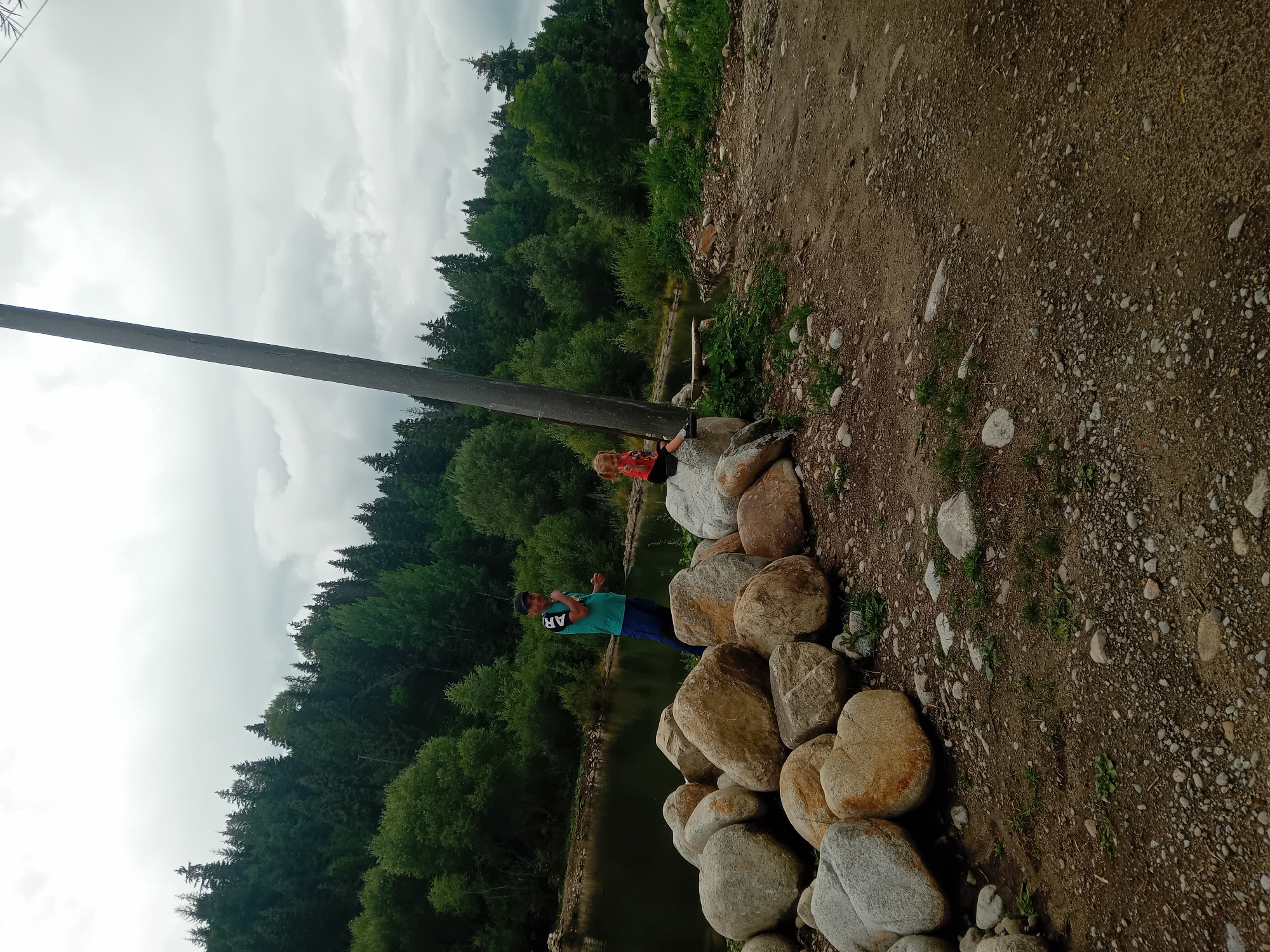 In Habovka, Slovakia, a man in a teal shirt and dark shorts stands on a pile of large, smooth river stones near the water's edge. He is looking towards the camera and has his left hand raised. To his right, a young girl with blonde pigtails sits on another large rock, wearing a red shirt. Behind them, a dark, calm body of water reflects the lush green trees and a cloudy sky. A tall, rough-hewn wooden pole stands diagonally in the foreground, angled towards the upper right of the frame. The ground is covered with dirt, gravel, and scattered smaller stones. The background is dominated by a dense forest of evergreen and deciduous trees.