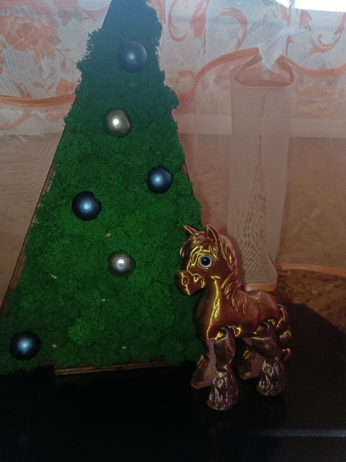 The scene is an indoor setting in Gorka, Russia, depicting two decorative objects arranged on a dark, reflective surface. To the left is a triangular, Christmas tree-shaped decoration, approximately 30-40 cm in height, covered in vibrant green, textured material resembling preserved moss. This decoration features five spherical ornaments: three dark blue and two silver-grey, distributed across its surface. To the right of the tree decoration stands a small figurine of a horse, approximately 15 cm tall. The horse figurine exhibits an iridescent or metallic finish, primarily reddish-brown with golden highlights, particularly on its raised features. It has prominent, round, light-colored eyes. The background consists of a window partially covered by two layers of curtains. The inner layer is a sheer, light-colored fabric, while the outer layer is a translucent, peach-toned curtain with a lighter, swirling floral pattern. No individuals are present, and no actions are occurring.