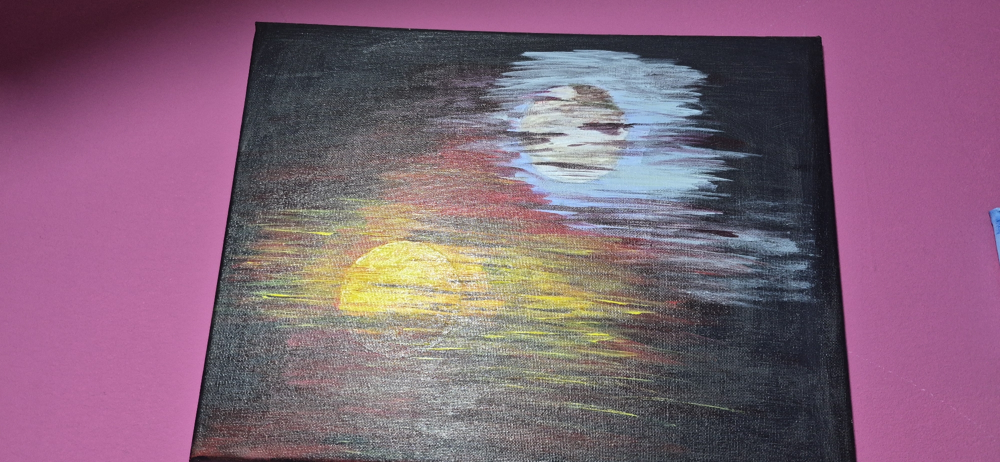 The image shows a painting of a sunset or sunrise over water. The sun is depicted as a bright yellow orb with streaks of yellow and orange radiating outwards. The water reflects the colors of the sky, with reds, oranges, and blues mixed in. Above the water, there is a hazy white and blue shape that could be clouds or a reflection of the moon. The painting is set against a dark background, possibly to emphasize the light from the sun. The painting is hanging on a magenta wall. The location in the city of Lom, Bulgaria is not discernible from the image.
