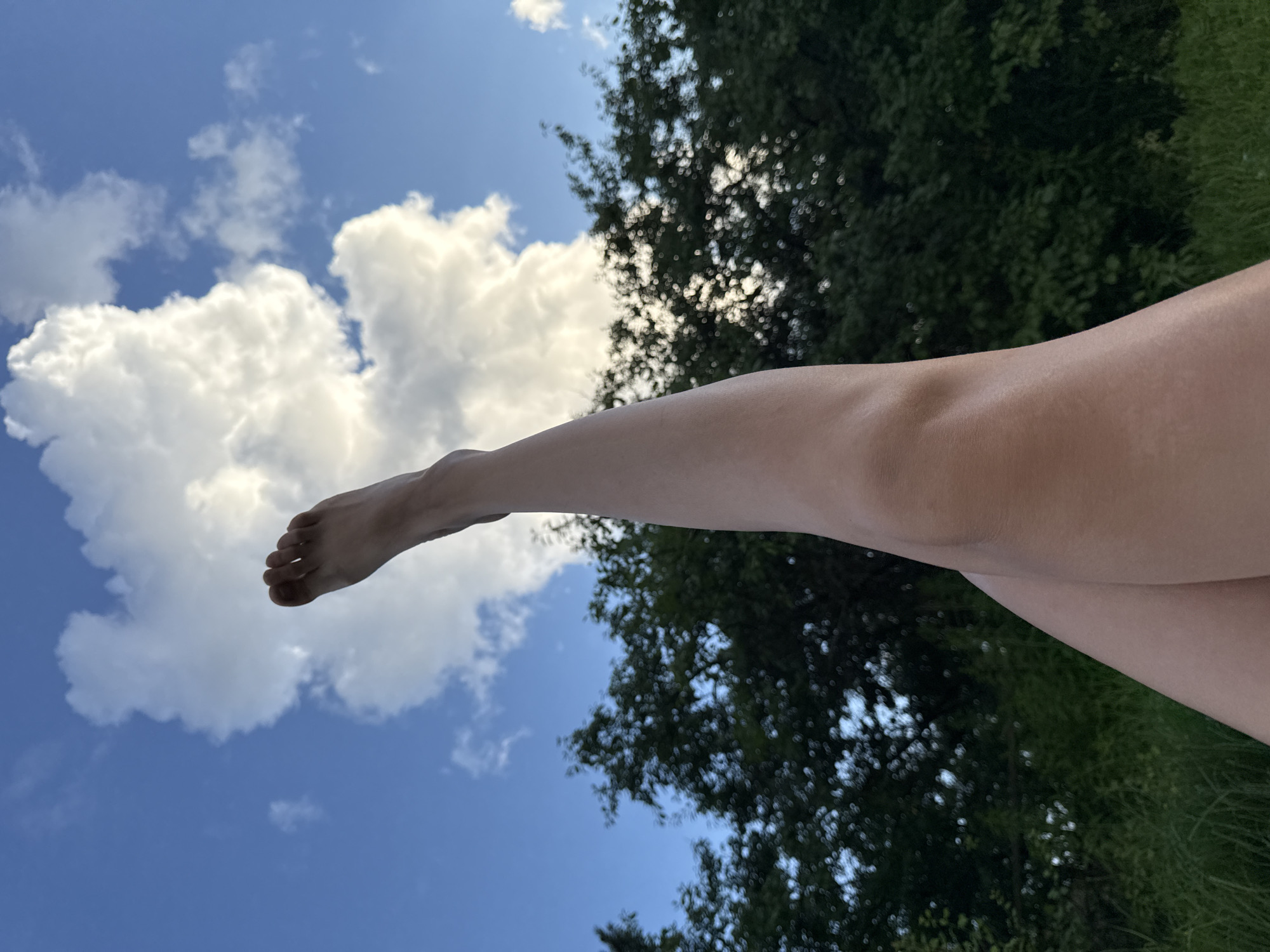 The image shows a person's legs extended upwards against a backdrop of a blue sky with white clouds and dark green foliage. The person's left leg is in the foreground, with the knee slightly bent and the foot pointing upwards towards the sky. The right leg is positioned behind and slightly below the left leg, with both legs creating an X-shape. The background is composed of trees and grass, suggesting an outdoor setting. The lighting indicates it is daytime. There are no discernible landmarks or elements that would specifically place the scene in the city of Ветлужский, Russia.
