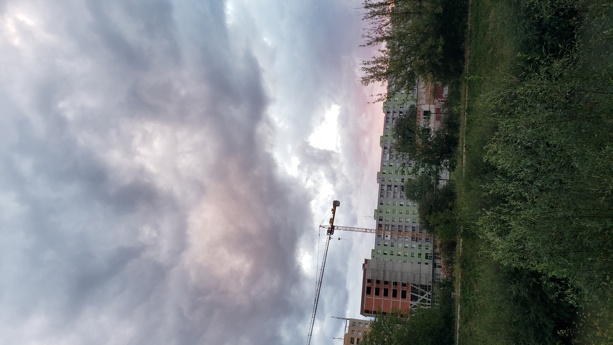 The image depicts a cloudy sky over a residential area with construction in progress, likely in the city of Prešov, Slovakia. A large crane is visible, extending over a building under construction that has a brick exterior and unfinished floors. Adjacent to this, a long, multi-story apartment building with light green siding and numerous windows runs horizontally across the frame. In the foreground, dense green foliage and trees create a natural barrier, suggesting the scene might be overlooking a park or a riverbank. The sky is filled with heavy, grey clouds, with hints of pink and orange light suggesting either dawn or dusk. There are no people visible in the image.