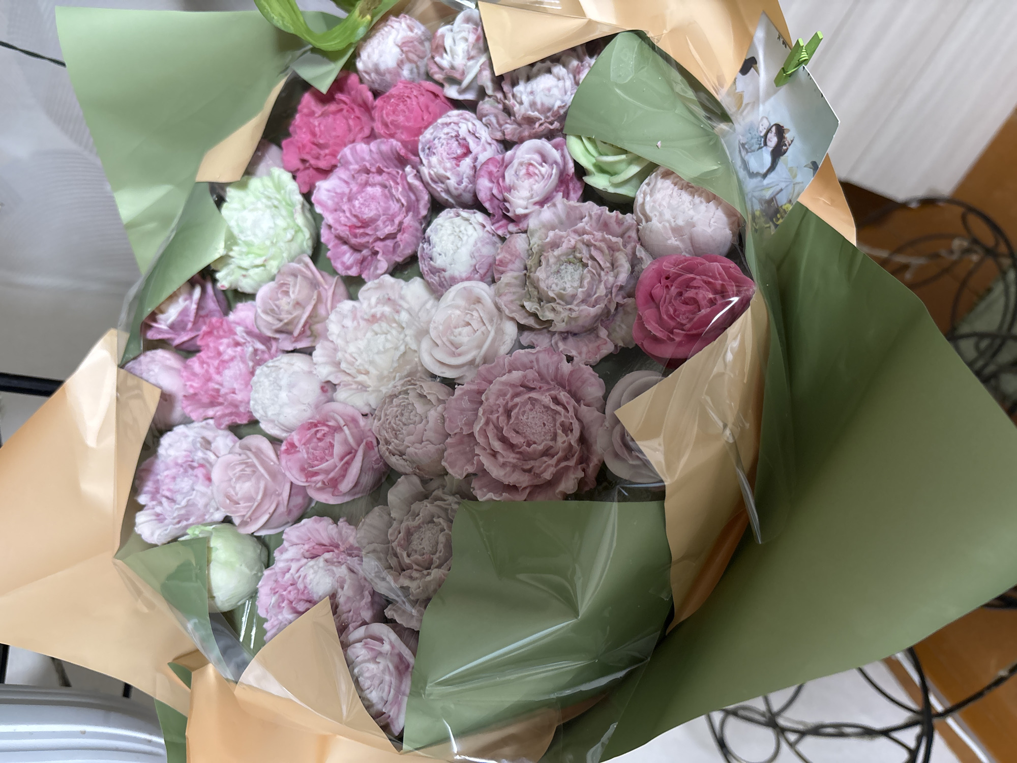 The image shows a close-up of a bouquet of soap flowers in various shades of pink, white, and green. The flowers are arranged in a bundle and wrapped in green and gold paper, with a clear plastic film covering the front. Some of the flowers resemble roses and peonies. In the lower right corner, a small printed card with an image of two people and a green clip is visible. The background is out of focus but appears to be an indoor setting with a white surface and some dark, wire-like structures. There are no people actively engaged in any actions within the frame. The specific location within Siberia, Russia, cannot be determined from the visual information provided.