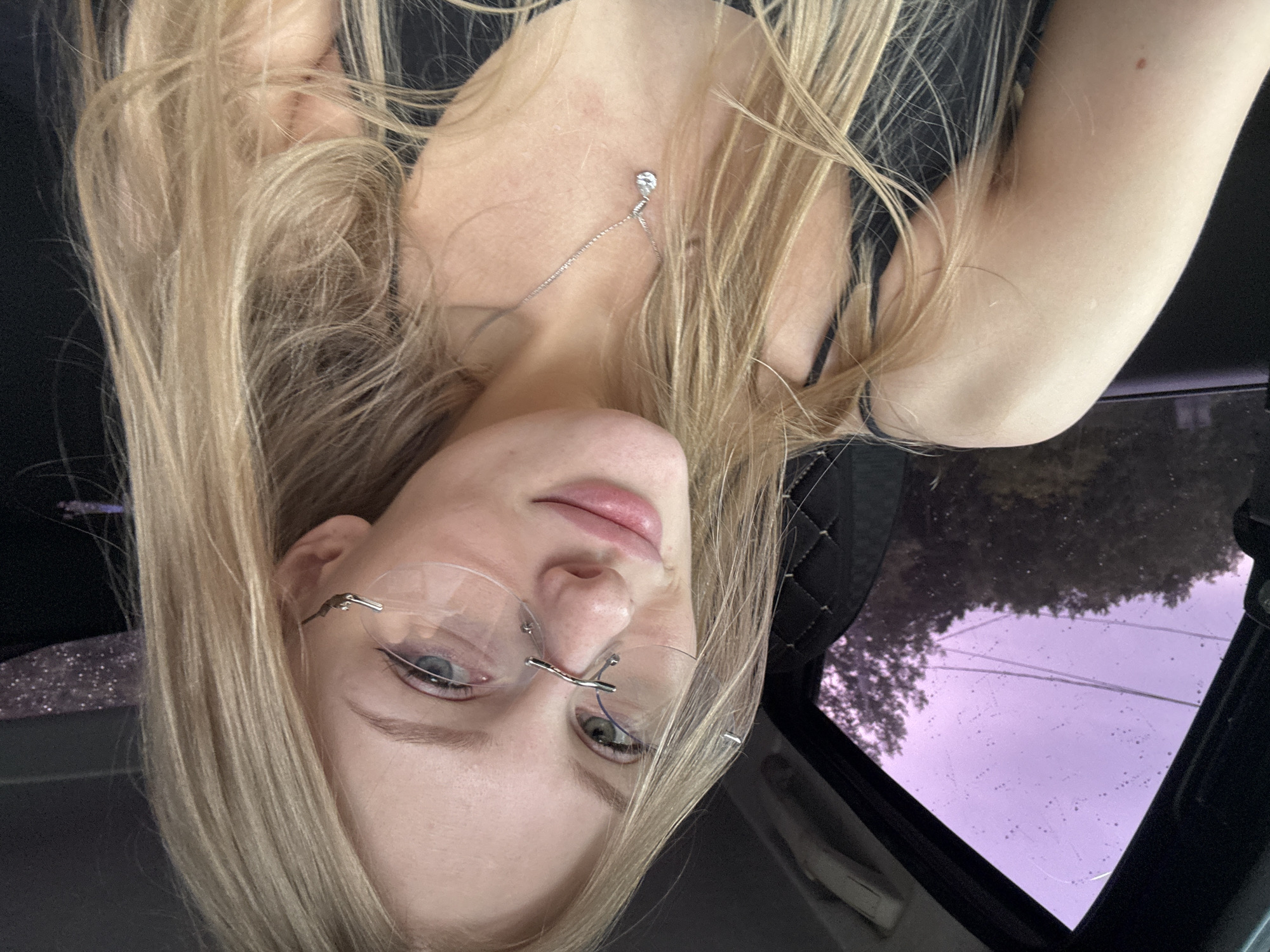 A young Caucasian woman with blonde hair, wearing glasses and a black camisole, is pictured inside a vehicle. She is looking towards the camera with a slight smile. The background shows trees and a purplish sky through the car window. The interior of the car, visible in the foreground, includes a black upholstered seat.