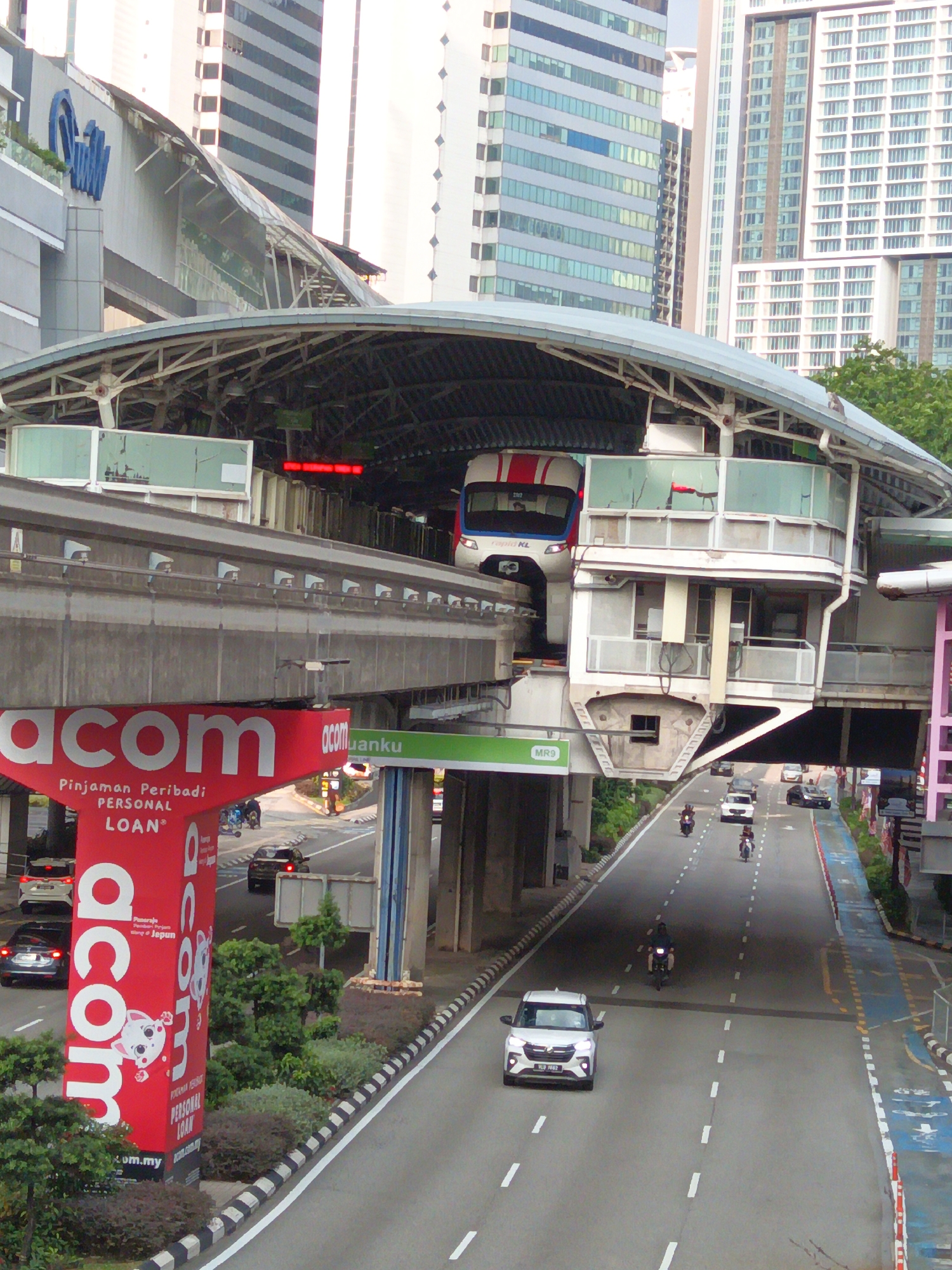This image captures a dynamic scene in Kuala Lumpur, Malaysia, likely from an elevated vantage point. The foreground is dominated by a large, red advertising pillar for "acom," a financial service offering "Pinjaman Peribadi PERSONAL LOAN." The pillar features stylized text and an illustration of a cartoon cat. To the left, a portion of a modern building with a blue logo is visible.
Above the pillar, a raised monorail track curves into a station structure. The station itself is a multi-level, modern design with glass and metal elements. A white and red monorail train is partially visible on the track within the station.
Below the monorail track and station, a multi-lane road runs through the urban landscape. Several vehicles are present, including white cars, a dark-colored sedan, and motorcycles. People are seen riding the motorcycles. Trees and greenery are interspersed along the roadside and in the background, suggesting a well-developed urban environment. In the distance, towering skyscrapers with modern architectural designs form the backdrop of the city. The overall impression is one of a busy, active city with a blend of transportation infrastructure, commercial advertising, and residential or office buildings.