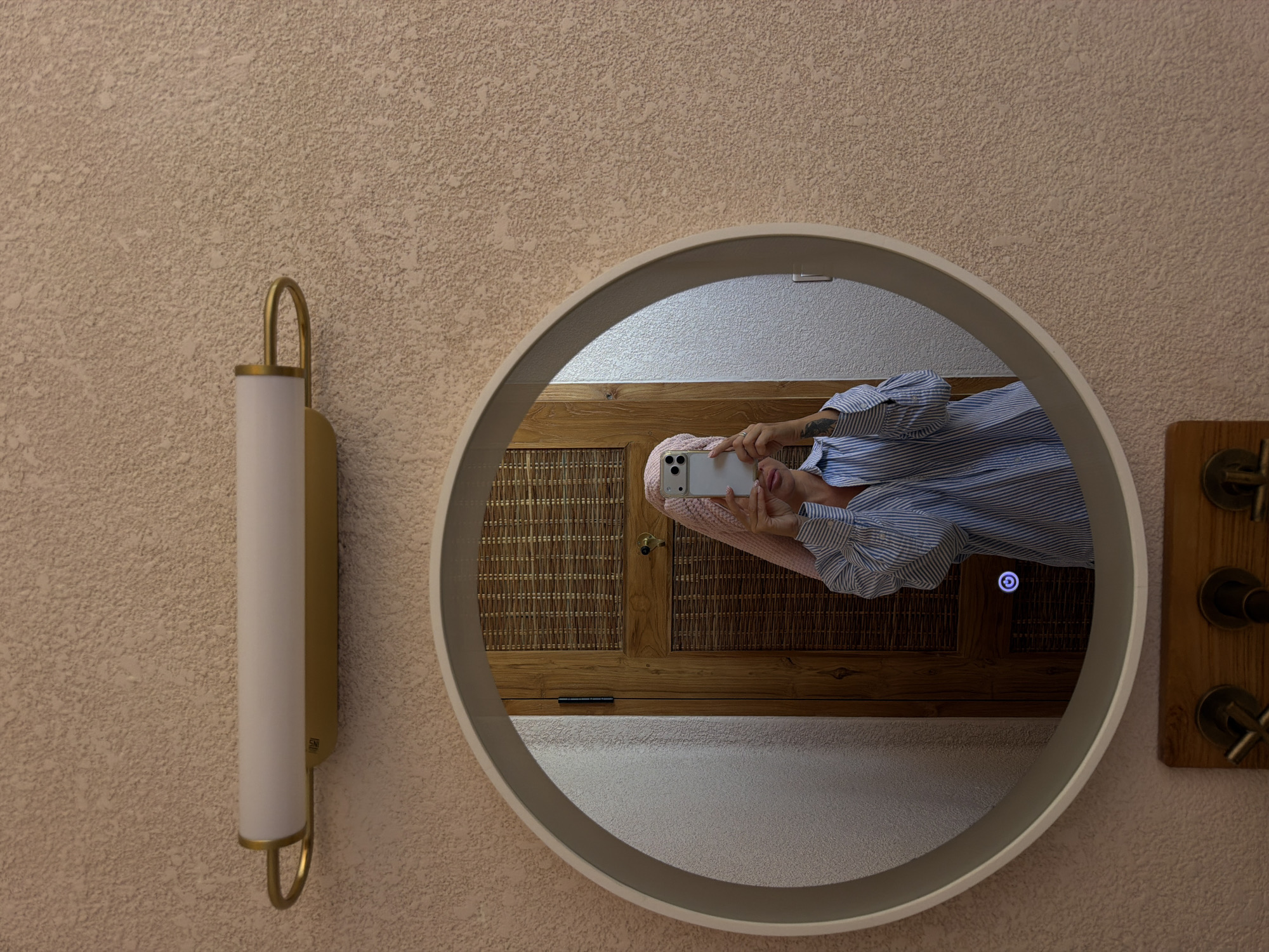 In a bathroom, a person with a pink towel wrapped around their head is taking a selfie in a round mirror. They are wearing a blue and white striped button-down shirt. The mirror reflects a wooden door with a woven pattern and a light fixture above it. The walls have a textured finish. The location is in Mengwi, Indonesia.