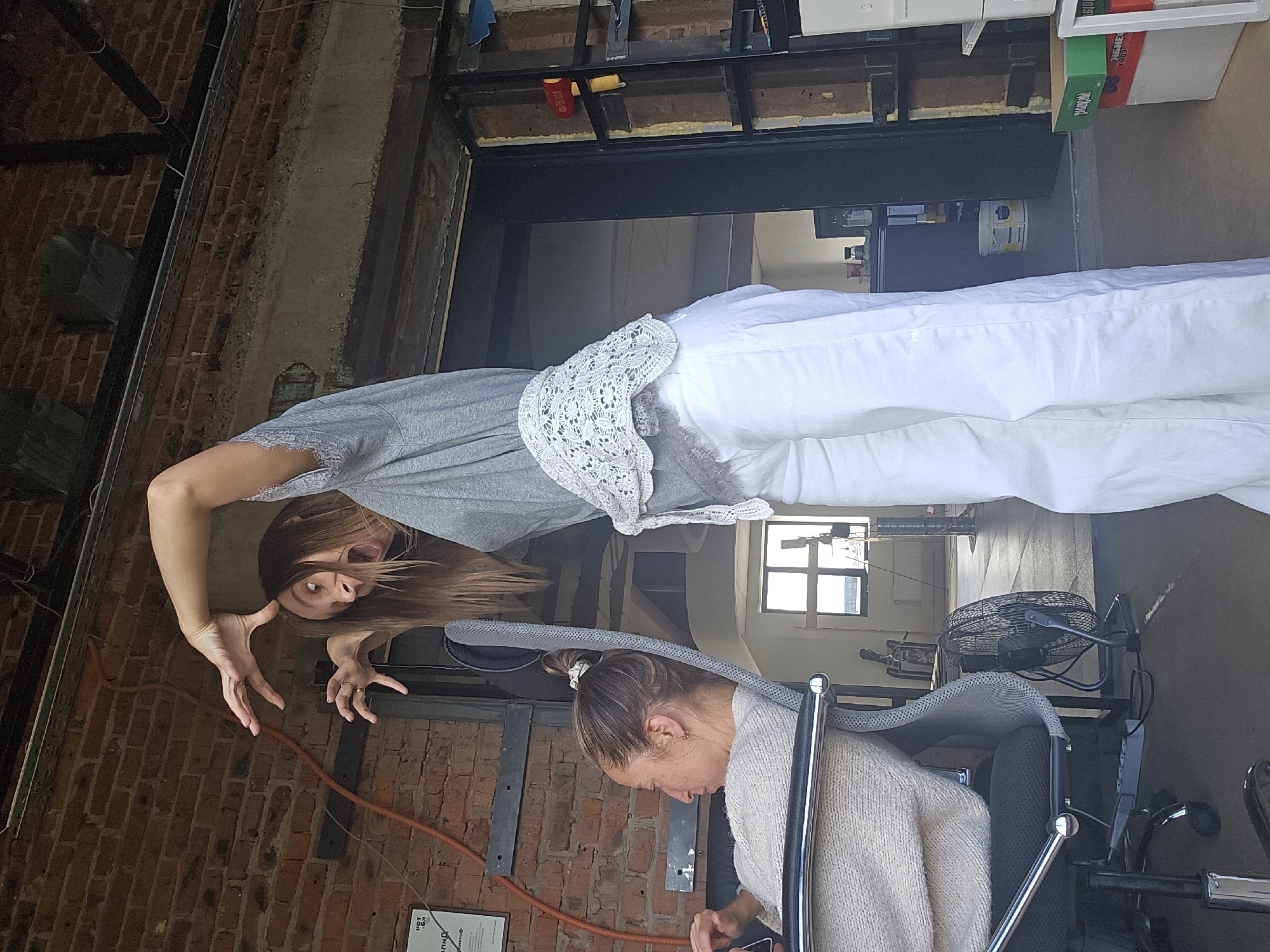 The image, correctly oriented by rotating it 90 degrees clockwise, captures an indoor scene, likely a casual studio or repurposed industrial space in Chaikovsky, Russia. The setting features exposed dark red brick walls and dark metal structural beams.
In the foreground, a woman with long brown hair, wearing a grey t-shirt with lace trim and white pants, strikes a playful, dramatic pose. Her arms are raised with fingers splayed, mouth open as if shouting or growling, and eyes wide, conveying an energetic and theatrical expression. A white lace garment is draped over her shoulder. To her right, a second woman with brown hair tied in a bun is seated in a black office chair. She wears a light-colored knitted sweater and is looking down, engrossed in a device held in her hands, likely a mobile phone.
The background includes various objects and structural elements. A large white object, possibly a mattress or cushion, is positioned near the first woman. Further back, a black electric fan on a stand is visible. Natural light enters the space through a distant window, indicating it is daytime. Miscellaneous items such as a red pouch or can (partially showing "RED" or "EDG") hang from a metal beam, and a green box with the partial label "Yoplait" can be seen among other containers. On the brick wall, a piece of paper displays Cyrillic text, including "ПРАВИЛА ТЕХНИКИ БЕЗОПАСНОСТИ" (Safety Rules) and fragments like "OT KE", possibly "HOT KEY".