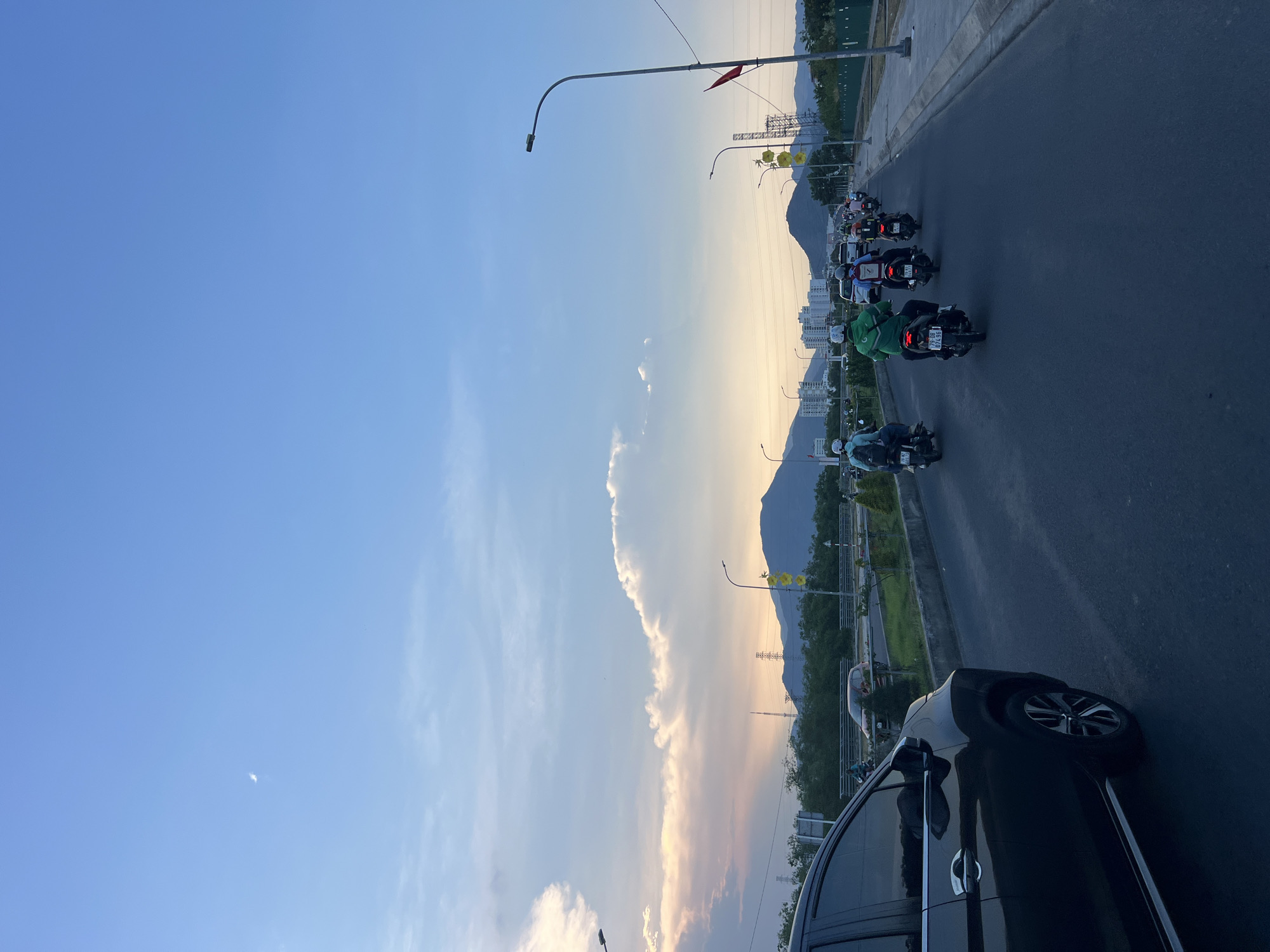 The image shows a street scene in Nam Nha Trang Ward, Vietnam. A line of motorbikes rides along a road, under streetlights. Mountains are in the background. A car is visible on the right side of the frame. The sky is blue with light clouds.