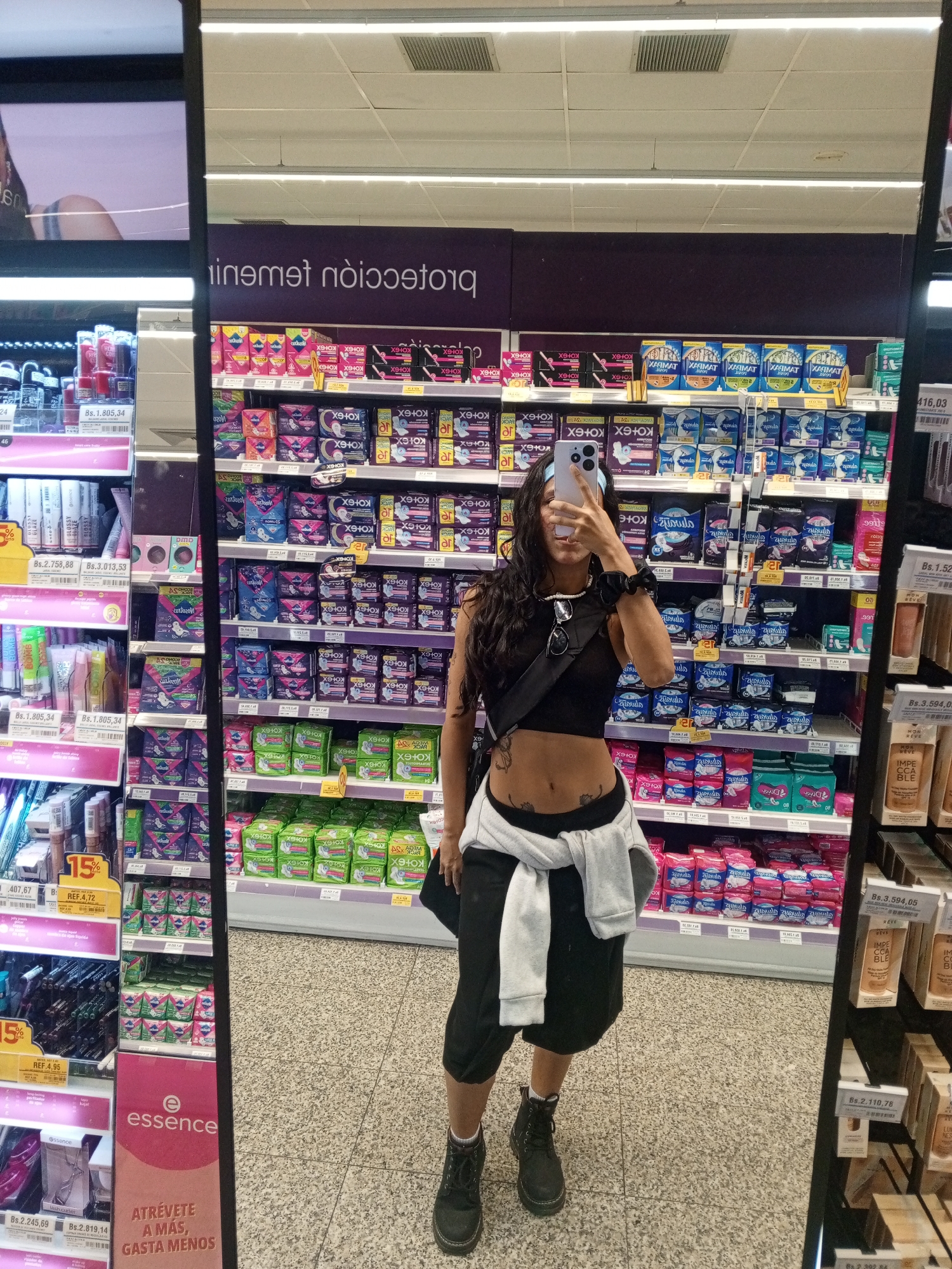 A person stands in a supermarket aisle, facing a mirror and taking a selfie with a smartphone. The person is wearing a black crop top, black shorts, and black boots. A light-colored sweater is tied around their waist. The aisle is stocked with feminine hygiene products, primarily Kotex brand items. Shelves on the opposite side of the aisle display cosmetics. Price tags in Bolivars (Bs) are visible on the shelves. The location is inferred to be a supermarket in Pampatar, Venezuela, based on the currency on the price tags.