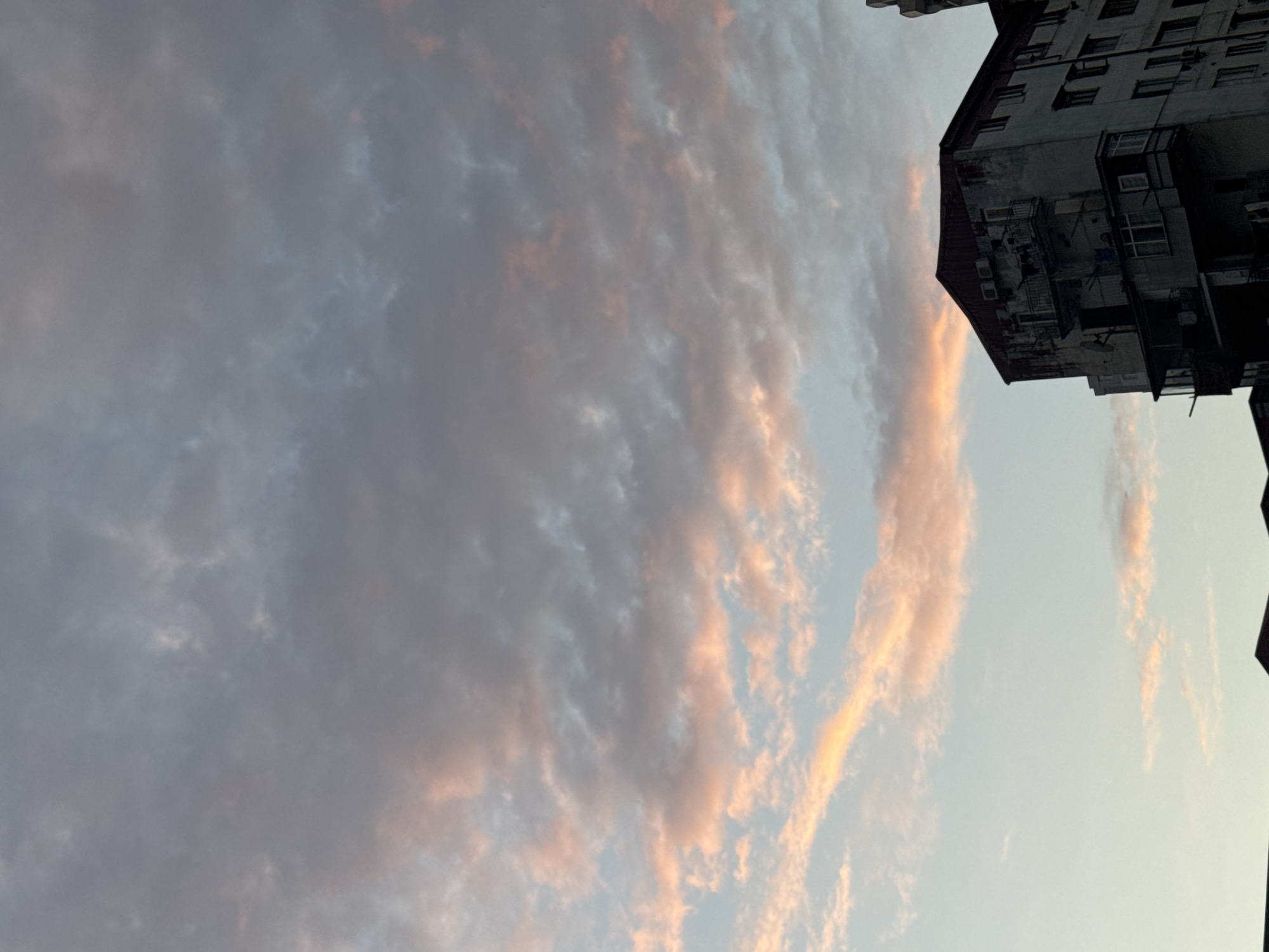 This is a vertical, medium-shot, eye-level photograph taken outdoors, likely in the city of Batumi, Georgia, showing the upper portion of an apartment building against a sky filled with clouds tinged with pink and orange hues, suggesting either sunrise or sunset. The apartment building is on the right side of the frame, dark and shadowed, with a weathered facade. Various architectural details are visible, including balconies, windows, air conditioning units, and satellite dishes. The sky dominates the upper two-thirds of the image, with a band of lighter, clear sky near the bottom, transitioning to a mottled expanse of gray and pinkish clouds above. No people or specific actions are discernible in the image. The overall mood is atmospheric, capturing a moment of natural beauty interacting with urban structures.