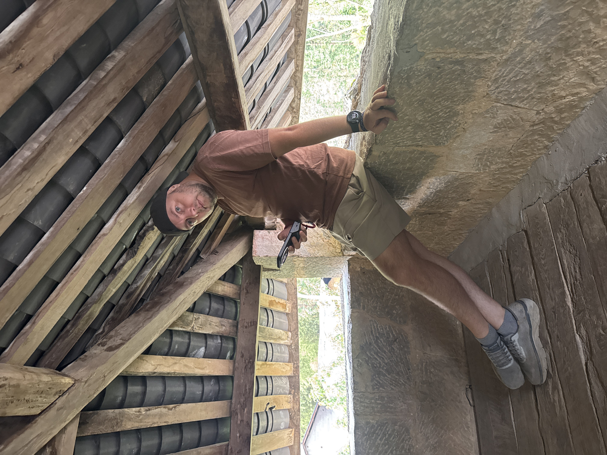 The image shows a man in Sà Phìn Commune, Vietnam, situated within an interior structure. He's wearing a brown t-shirt, khaki shorts, grey socks and shoes, and a black cap. He is holding a small black device in his left hand and is propping himself up against a rough stone or concrete wall with his right hand. He is wearing a large black watch on his left wrist.
To his left, wooden beams and a tiled roof can be seen. Through an opening or window, greenery is visible in the background. The ground appears to be made of wood planks or stone. The overall impression is that the man is exploring or inspecting an old structure.