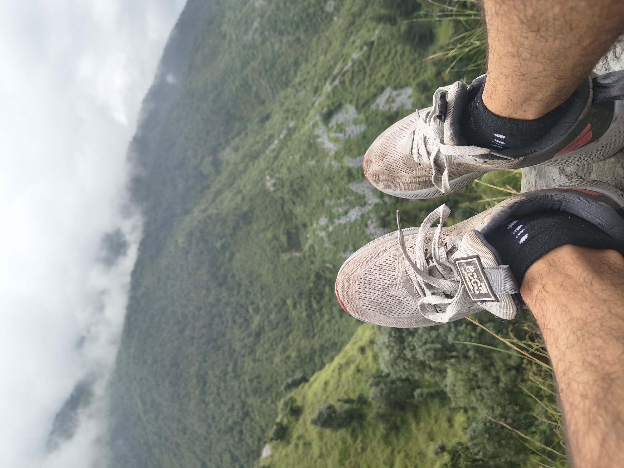 A first-person perspective shot shows a pair of dirty, light-colored athletic shoes with grey laces, worn with black socks, resting on a stone. The shoes are positioned in the foreground with their toes pointing upwards and slightly outwards. Behind the shoes, a lush, green mountainside slopes downwards, covered in dense vegetation and trees. The upper part of the image is dominated by a cloudy, overcast sky that partially obscures the higher peaks of the mountains. The scene suggests a hike or a break during outdoor activity in a natural, mountainous environment.