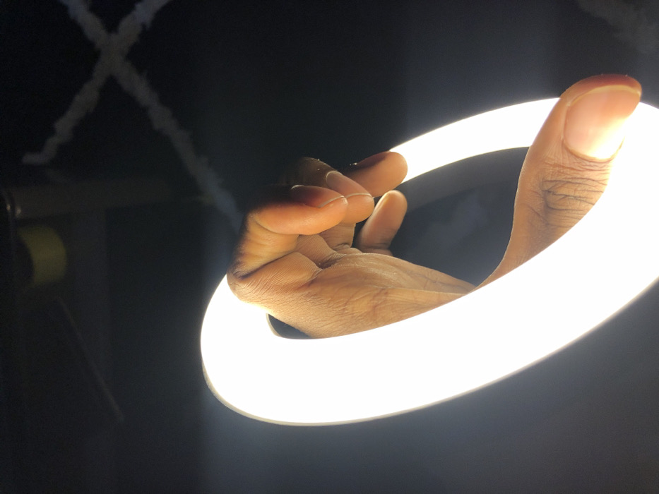 The image, captured in Tema, Ghana, depicts a close-up, upward-angled view of a human right hand actively holding or manipulating a brightly illuminated, C-shaped light source, consistent with a segment of an LED ring light. The hand, displaying dark brown skin pigmentation, has its fingers curled around the upper arc of the light, with the thumb positioned along the lower arc. The light emits a strong, continuous white glow, creating high contrast and illuminating the skin texture. The background is largely dark and out of focus. Faint environmental elements are discernible in the upper frame: a blurry yellow object and a thin dark linear element extend from the upper left, while two diffuse, white linear elements form an 'X' pattern in the upper right.