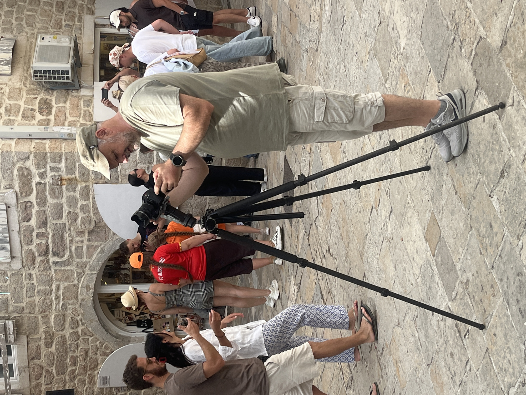 A diverse group of people gathers on a paved outdoor area, possibly a pedestrian street or square, under bright sunlight. The ground is made of light-colored, irregularly shaped stone pavers, and stone buildings line the left side of the scene.
In the center foreground, a man with a beard and a baseball cap is bent over, adjusting a black camera tripod. He wears a light-colored t-shirt, khaki cargo shorts, light sneakers, and a dark smartwatch on his left wrist. Behind the tripod, another person holds a camera with a large lens. Further back, several individuals are visible, including a person in a white t-shirt and light blue jeans, and another in dark clothing.
To the right of the tripod, a woman wearing a red t-shirt with "FC AUGSBURG" printed on the back stands, her braided hair visible. Nearby, another person in a plaid dress and a straw hat is looking towards a shop window. In the immediate foreground to the right, a man with a beard and a woman with long dark hair are conversing or looking in the same direction. The woman is dressed in patterned pants and flip-flops, while the man wears a brown t-shirt and light-colored shorts.
On the left side of the image, a stone wall features an outdoor air conditioning unit and some framed artwork. The background reveals glimpses of shop windows with items displayed. The overall activity suggests a casual public gathering, likely tourists or locals, engaging in sightseeing or photography. The bright lighting indicates a sunny day. No strong emotions are distinctly visible; most individuals appear relaxed and observant.