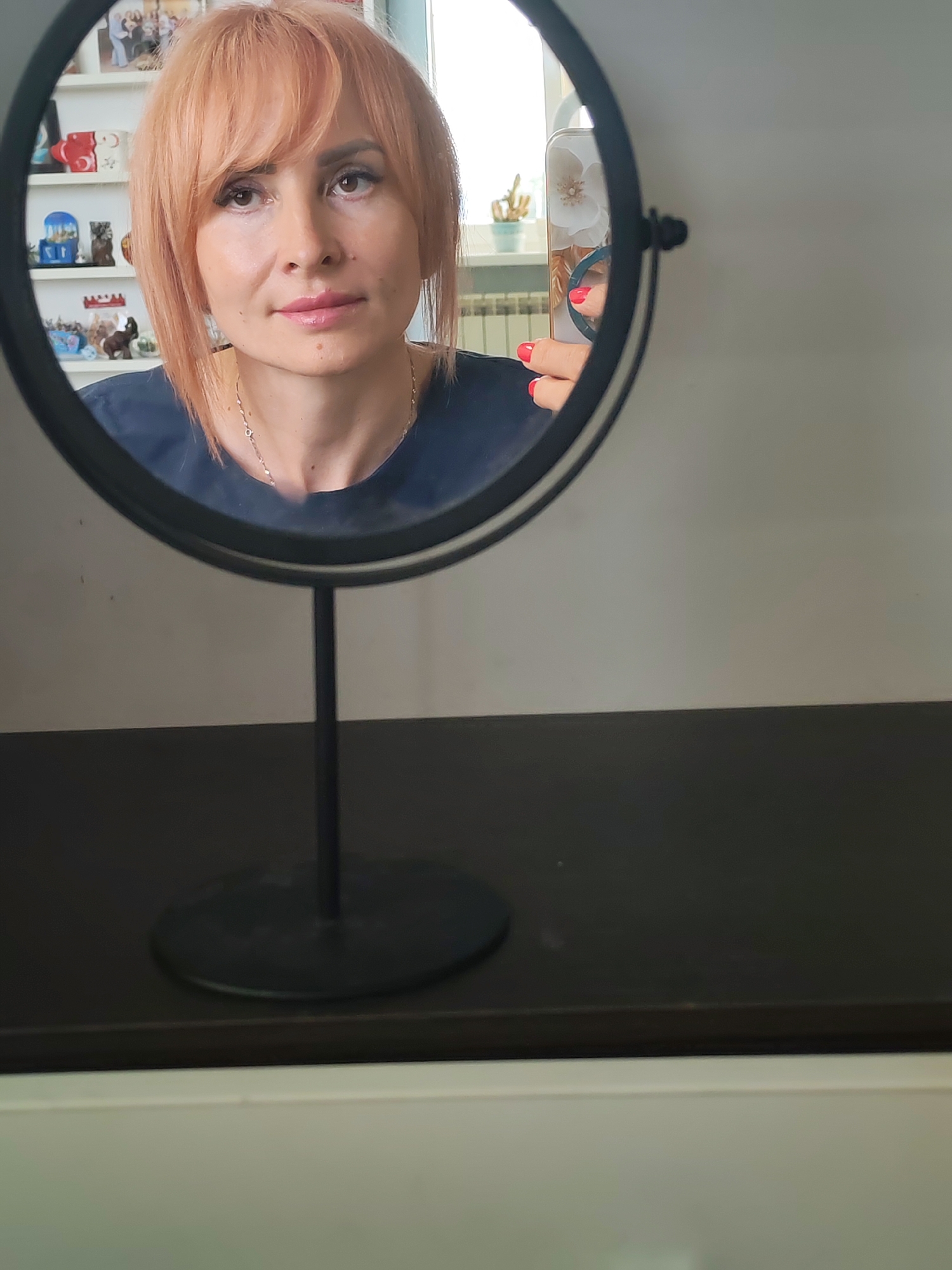 An overhead shot shows a woman looking into a black framed mirror. The woman has short, light orange hair and fair skin. She is wearing a dark blue top and a delicate silver necklace. Her nails are painted bright red. The mirror reflects her face and upper body, and also shows a phone in her right hand. In the background of the mirror's reflection, shelves filled with knick-knacks are visible, as well as a window with a radiator and plants. The mirror is positioned on a dark surface, possibly a dresser or table, in a room. The overall lighting is natural and soft.