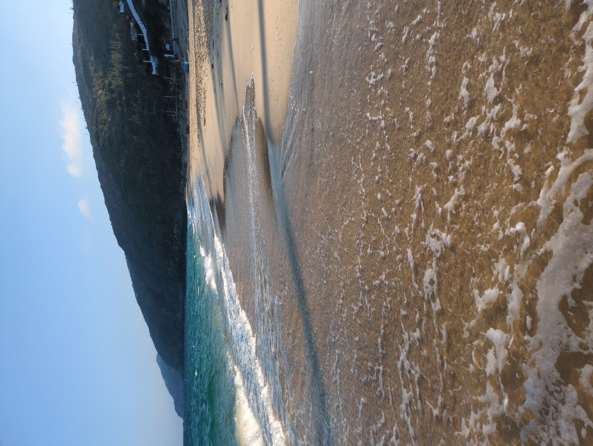 The image depicts a coastal scene at East Quy Nhon Ward, Vietnam. In the foreground, foamy white waves from the turquoise-green ocean recede across light brown, wet sand, creating intricate patterns. The midground features a wide, light brown sandy beach, illuminated by sunlight casting long shadows from an unseen source to the left. On the dry sand closer to the hillside, a small, light blue boat is visible, along with a darker, possibly red, boat further back near the base of the hillside. Several dark, elongated shadows are cast across the sand. The background is dominated by a densely vegetated, dark green mountain range sloping steeply upwards, extending along the coastline into the distance. White structures, possibly buildings or retaining walls, are visible built into the lower slopes of the hillside, interspersed with the vegetation. A wooden structure, resembling a raised platform or small pier, is situated at the edge of the sand near the base of the mountain. The sky above is clear and light blue, with a few faint wisps of white cloud visible. No individuals are present in the scene.