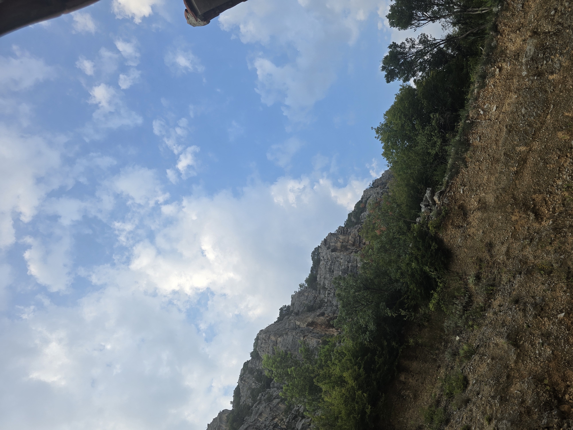 The image depicts a steep, rocky hillside covered with lush green trees and vegetation. The sky above is a vibrant blue, dotted with scattered white clouds. In the upper right corner, a portion of a building's roof is visible, featuring red tiles and concrete structure. The foreground of the image is dominated by the rugged terrain of the hillside, with loose rocks and soil visible. The overall scene suggests a natural, outdoor environment, possibly in a mountainous or hilly region. Based on the context provided, this scene is located in Podgorica, Montenegro.
