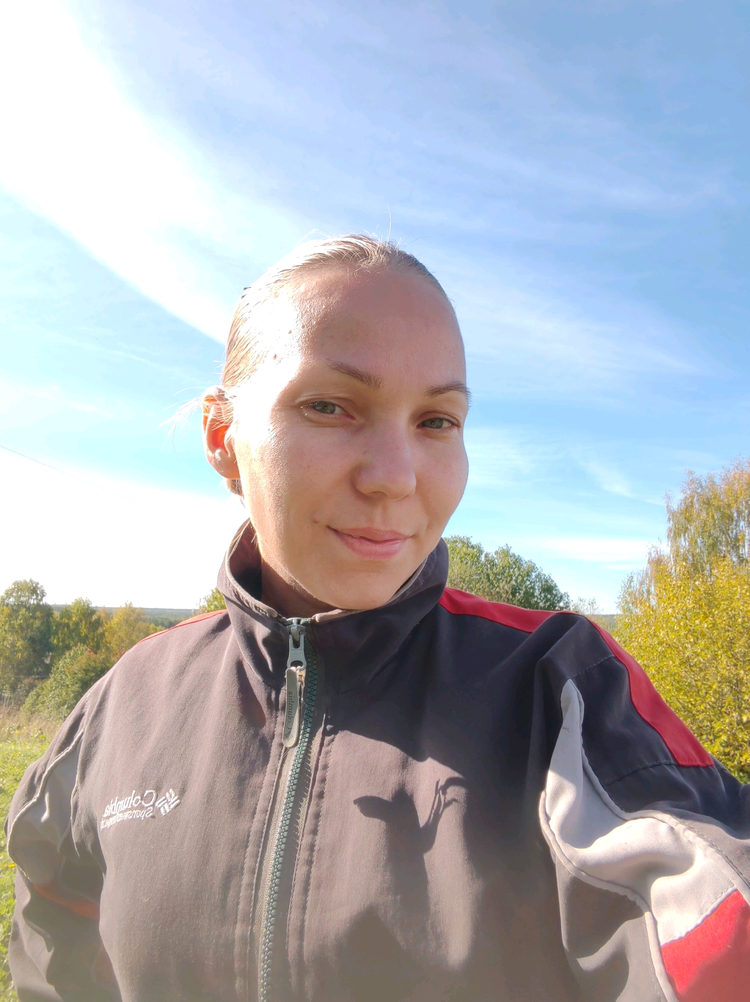 A young woman is captured in a close-up, outdoor selfie against a backdrop of a bright blue sky with wispy clouds and a distant landscape of trees. She is wearing a dark, zippered jacket with red and white accents. The sunlight illuminates her face, highlighting her fair skin and light-colored hair pulled back. Her gaze is directed towards the camera with a slight smile. The image appears to be taken in a rural or natural setting, possibly outside of Chuchlem, Russia, given the natural light and vegetation.