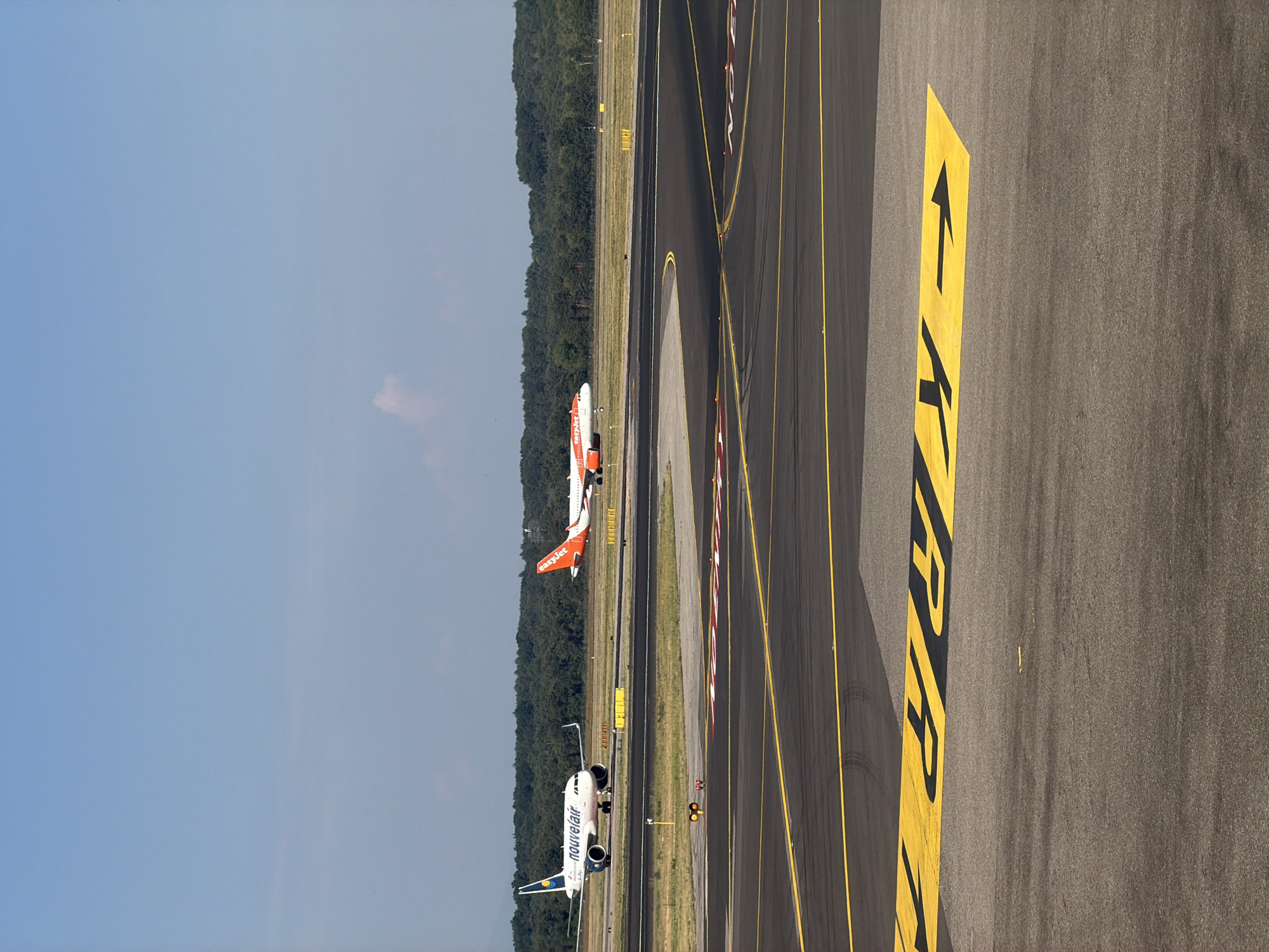 This image captures an outdoor scene at Malpensa Airport in Ferno, Italy, on a clear, sunny day. The foreground features an asphalt runway with yellow directional arrows and markings painted on its surface. In the mid-ground, two commercial airplanes are visible on separate taxiways. The airplane on the left is white with blue and yellow accents and the word "nouvelair" emblazoned on its side. The airplane on the right is white with prominent orange branding, identifiable as "easyJet". Both aircraft appear to be stationary or taxiing at a moderate speed. The background is dominated by a dense line of green trees, suggesting a wooded area bordering the airport. The sky is a bright, clear blue with a few faint clouds. The lighting indicates it is daytime, with sunlight casting shadows on the runway. There are no visible people in the frame.