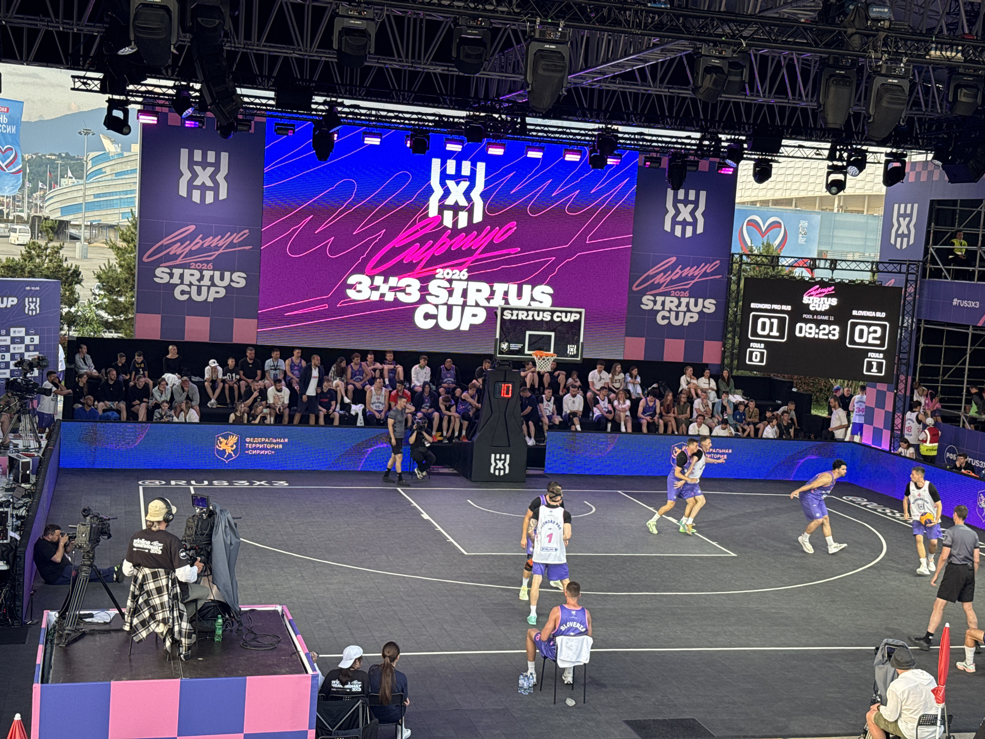 The image shows a 3x3 basketball game in progress on an outdoor court in Sirius, Russia. Spectators are seated in the background, and large screens display event branding and live scores. The scoreboard shows "01 09:23 02," indicating the teams and the current game time. Players in purple and white uniforms are on the court, with one player in a white jersey dribbling a basketball. Camera operators are positioned around the court, filming the event. The overall scene depicts a professional sports event with a focus on the 3x3 basketball competition.