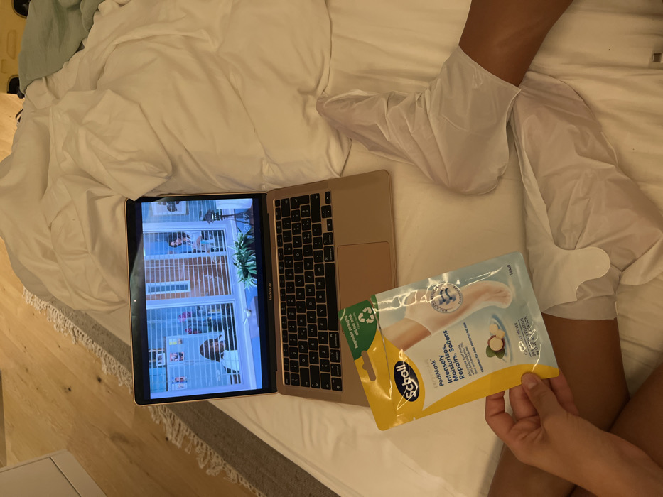 In Vila Nova de Cerveira, Portugal, an indoor scene depicts a person engaged in self-care and entertainment. A gold Apple MacBook Air laptop rests open on white, rumpled bedding, displaying an interior scene from a video featuring multiple individuals and window blinds. The person's lower legs and feet, with a tanned complexion, are visible. Both feet are encased in white, single-use foot masks. The person's left hand holds a yellow and light blue package of "Scholl PediMask Intensely Moisturises, Repairs, Softens," which indicates "1 Pair" and contains avocado oil. A light-colored wooden floor and a textured gray rug are visible in the background to the left.