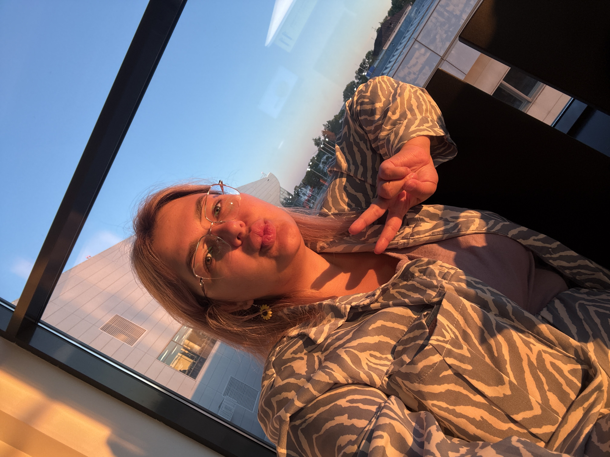 A young woman with blonde hair and glasses is taking a selfie in front of a large window with a city view. She is wearing a grey and white zebra-print jacket over a light purple t-shirt and is making a peace sign with her right hand while puckering her lips. The sun is setting, casting a warm orange glow on the side of a building visible through the window, and the sky is a clear blue. The scene appears to be indoors, possibly in an office building or a modern apartment, with dark furniture in the foreground. The location is identified as the city of Khabarovsk, Russia.