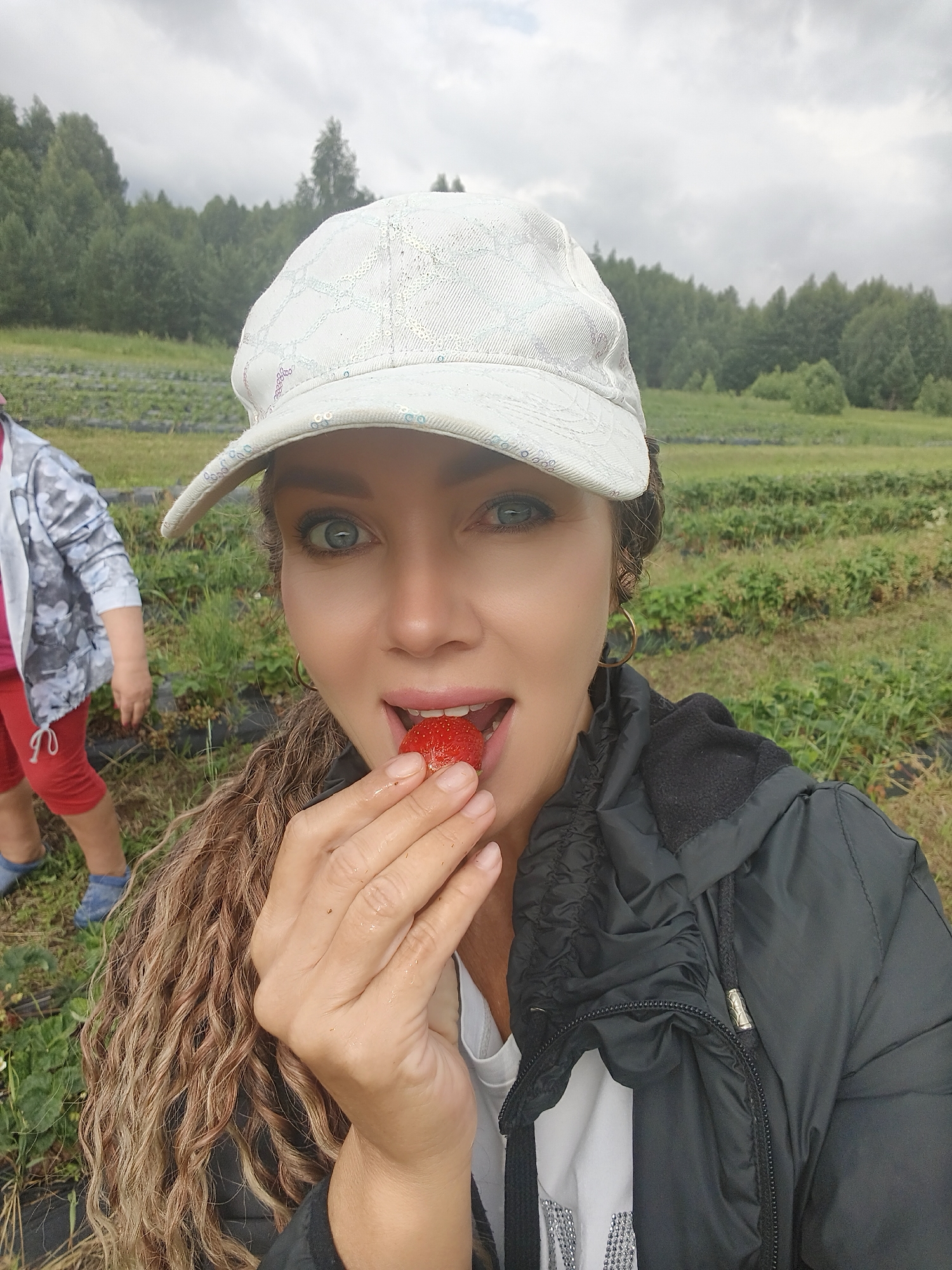 A woman is shown in a selfie, with a strawberry held to her mouth, at a strawberry field under an overcast sky. She is wearing a white baseball cap with a patterned design, a black jacket over a white shirt, and large hoop earrings. Her right hand is holding the strawberry and is near her face. To the left of the woman, a child is visible from the waist down, wearing red shorts and blue shoes. The background consists of rows of strawberry plants and a distant treeline. The location is an outdoor strawberry field.