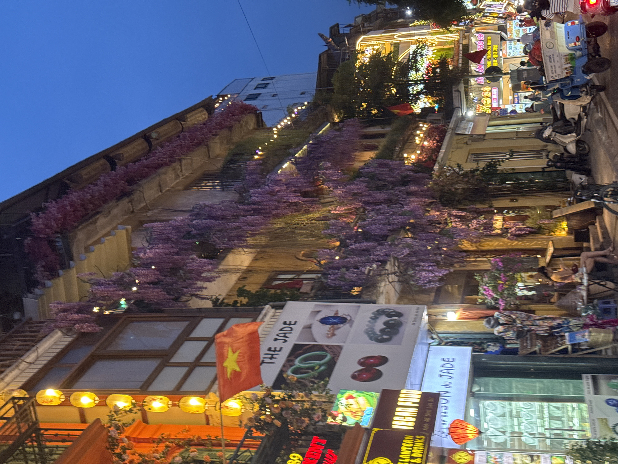 This image captures a vibrant street scene at dusk in Hà Nội, Vietnam, likely in a popular tourist area such as the Old Quarter. The architecture features multi-story buildings with balconies adorned with cascading purple wisteria flowers. Strings of warm, yellow fairy lights are draped along the building facades, illuminating the street below.
Several shops line the street, indicated by illuminated signs. One prominent sign reads "THE JADE" and displays images of jewelry. Another sign, partially obscured, appears to advertise "VEGAN FOOD." A red Vietnamese flag is visible, adding to the local atmosphere.
The street is bustling with activity. Various motorbikes and scooters are parked or in motion. A three-wheeled motorized cart, laden with goods, is a focal point in the foreground. Several people are seen on the street, some walking, others sitting at tables outside a shop. One woman is seated at a small table, looking down, possibly at her phone. The lighting suggests it is early evening, with the sky a deep blue and the artificial lights of the shops and streetlights creating a warm, inviting glow. The overall impression is one of a lively and picturesque urban environment.