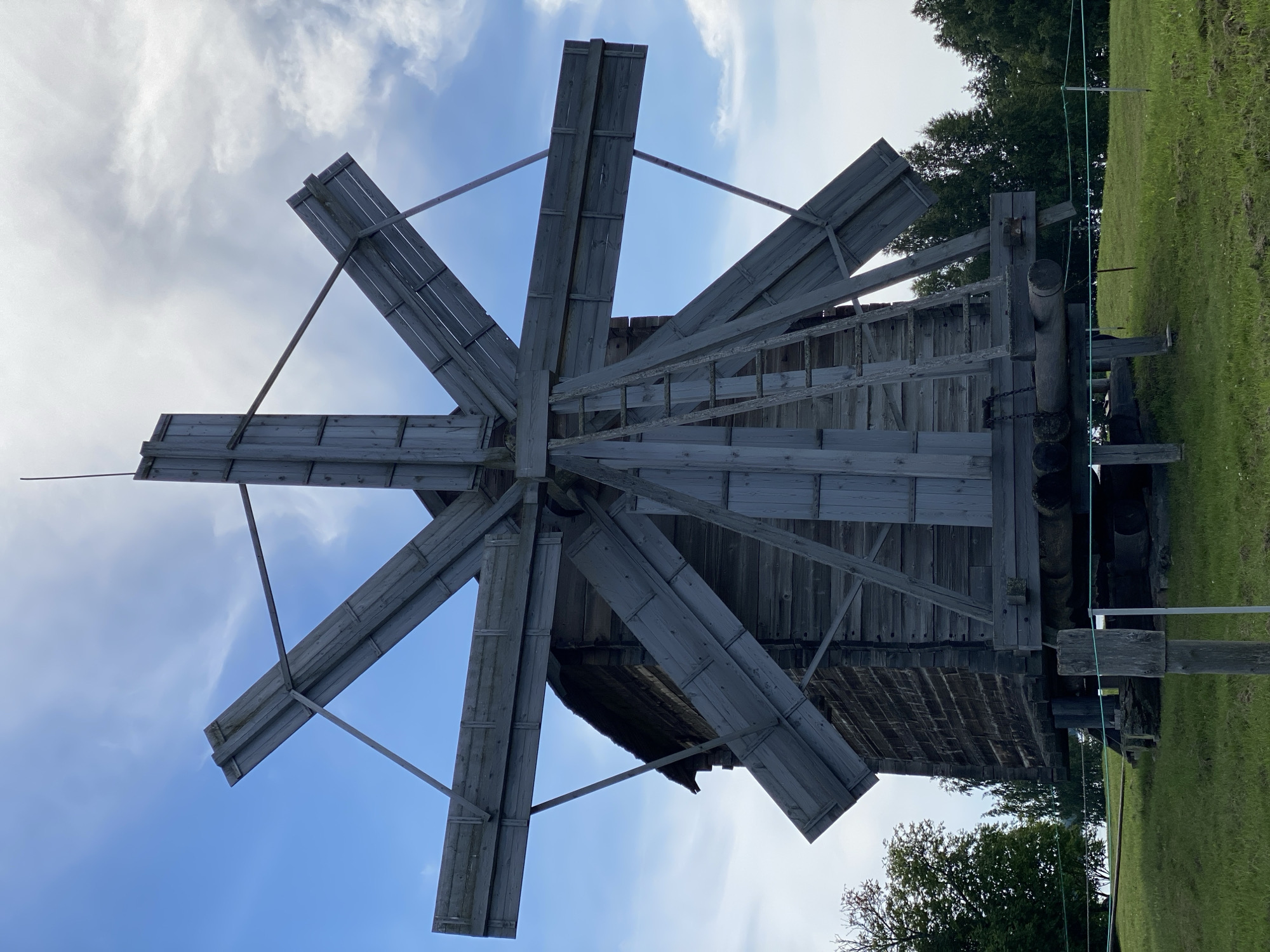 The image shows a traditional wooden windmill with six large, weathered blades, tilted upwards against a bright blue sky with scattered clouds. The windmill is constructed of dark, aged wood, with vertical planks forming the body of the structure. A wooden ladder is attached to the side of the windmill, leading up to the base of the blades. The windmill sits on a grassy hill, with lush green grass in the foreground. In the background, there are trees and a body of water, possibly a lake or river. The overall scene suggests a rural, historical setting, likely in the village of Boyarshchina, Russia, known for its wooden architecture and natural beauty. There are no people visible in the image. The lighting indicates it is daytime, likely late morning or early afternoon, with the sun high in the sky. The composition focuses on the imposing structure of the windmill, highlighting its rustic charm and engineering.