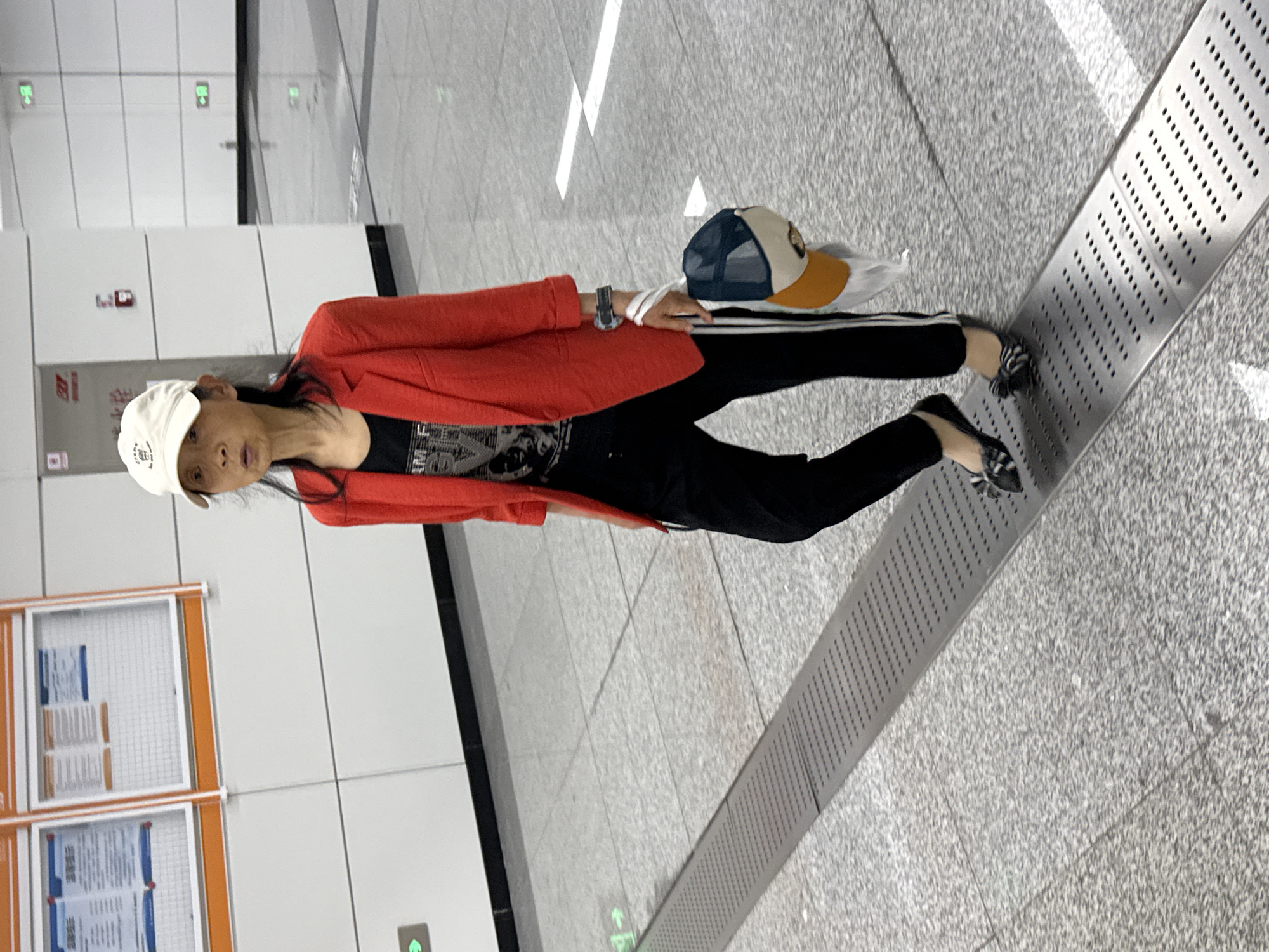A person wearing a white baseball cap, red blazer, black top, and black pants stands in what appears to be a subway station. They are holding a second baseball cap with tan and blue accents in their right hand. The floor is tiled gray with linear metal grates running diagonally across the scene. A wall is visible with various panels and signs. This is potentially the Liangjiang, China area based on the prompt.