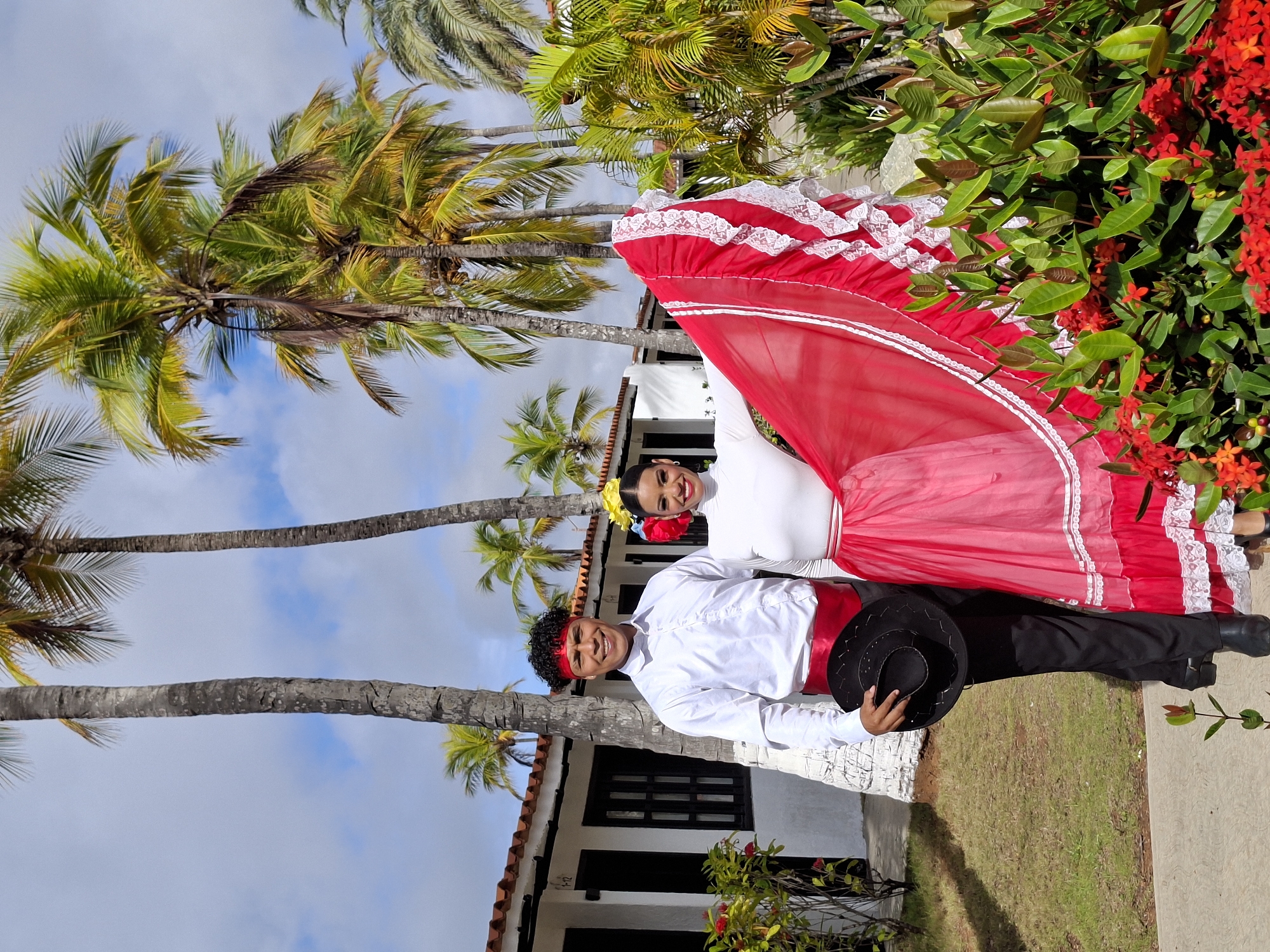 A man and a woman, dressed in traditional vibrant attire, pose for a photograph outdoors amidst lush tropical foliage and palm trees. The man, with curly dark hair and a red headband, wears a white collared shirt, black pants, and a red sash, holding a black hat in his right hand. The woman, with dark hair adorned with a yellow and red flower, wears a white turtleneck top and a voluminous red and pink layered skirt with white lace trim. They stand to the left of a white building with orange tiled roofs, likely a hotel or resort. Red ixora flowers are in bloom in the foreground, adding to the colorful scene. The sky above is partly cloudy, with bright sunlight illuminating the tropical environment. This scene is consistent with the tourist-friendly areas of Porlamar, Venezuela, known for its beaches and resort accommodations.