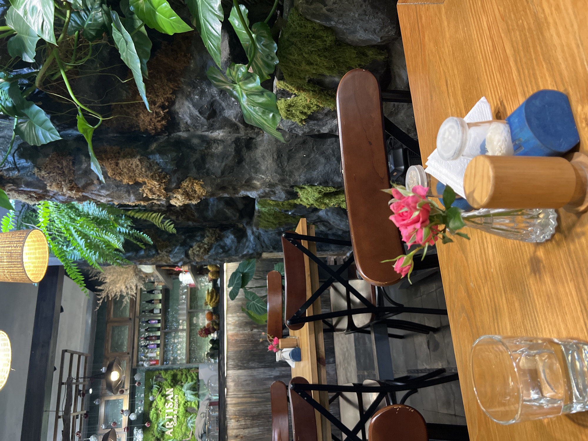 The image shows a section of the Artisan Cafe & Eatery in Nha Trang, Vietnam. The scene is indoors, featuring a wooden table setting with brown wooden chairs. On the table, there is a small vase of pink roses, a napkin holder with napkins, and a glass. The background includes a rock wall covered in moss and greenery. Beyond the wall, there is a view into another area of the cafe, showing wine bottles on shelves and the cafe's signage. The lighting suggests a relaxed atmosphere. There are no people visible in this close-up section of the cafe.