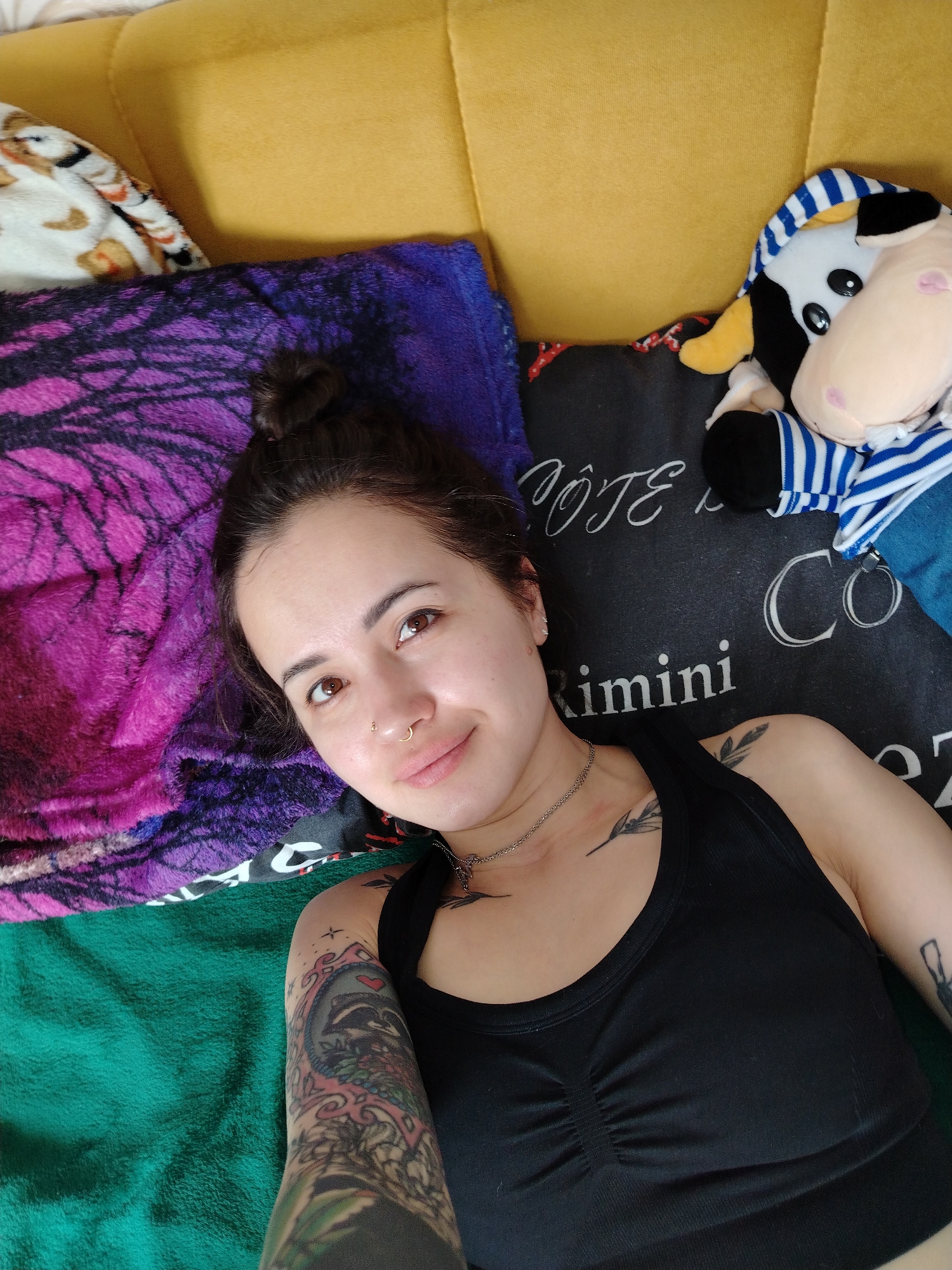 An overhead shot shows a female individual lying prone on what appears to be a bed or sofa in an indoor residential setting within the city of Unknown, Unknown. The individual is centered, gazing directly at the camera with a neutral expression, exhibiting a slight upward curve to the lips. Her dark brown hair is secured in a bun, visible at the top of her head. She presents with brown eyes and two facial piercings: a small gold hoop in her left nostril and a small gold stud in her right nostril. She wears a thin silver chain necklace and a black ribbed sports bra or tank top. Distinct tattoos are visible on her left arm, forming a colorful full sleeve that includes a framed image of a raccoon with a red heart, and a smaller, dark, plant-like design on her right collarbone/shoulder area.
The immediate environment includes a yellow upholstered surface serving as a headboard or backrest behind her. To her left, a purple fuzzy blanket with a dark, intricate pattern resembling branches is visible. Beneath her left arm and extending to the lower left, a green fuzzy blanket is present. To her right, a black rectangular pillow with partially visible white text, including "ÔTE", "Rimini", and "Co", supports a stuffed cow toy. The cow toy is black and white, featuring a pink snout, yellow horns, and is dressed in a blue and white striped top and blue overalls. A white blanket with a light brown animal print pattern is partially visible in the upper left corner of the frame.