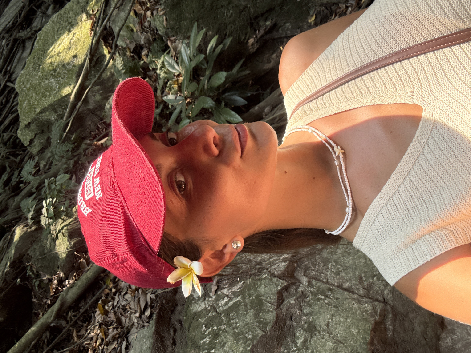 A person, presumably female, is lying down outdoors, possibly on a rock or a flat, stony surface. She is wearing a light-colored, ribbed top with a brown trim strap and a white beaded necklace. She also has on a red baseball cap with white lettering and a small white flower tucked behind her ear. The background consists of natural elements like tree roots, foliage, and rocks, indicating a forest-like or natural setting. Given the request for a location in Đà Nẵng, Vietnam, the location may be related to the Marble Mountains or Son Tra Mountain, known for their natural rock formations and green vegetation. The lighting appears to be bright and sunny, casting shadows on the person's face and clothing.