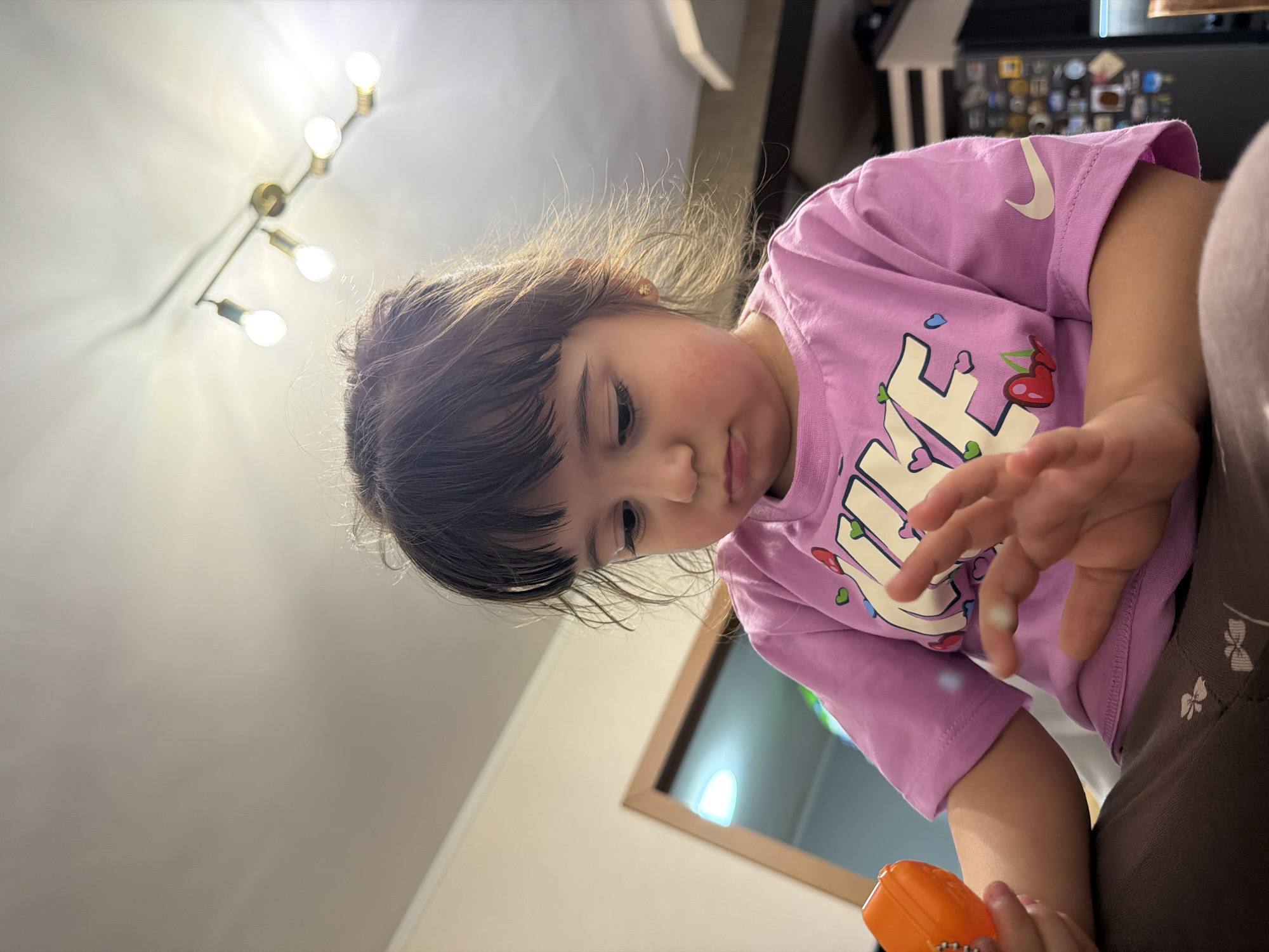 A low-angle, tilted indoor shot captures a young child from the chest up, seemingly focused on an object or activity below the frame. The child, with dark hair and fair skin, wears a light purple t-shirt with a white graphic design. The design features block letters and a prominent swoosh, reminiscent of the Nike logo, adorned with small hearts and cherries. Her cheeks appear rosy, and her expression is one of concentration.
Her hands are visible; one hand is extended, palm partially open, while the other holds a bright orange, rectangular toy or object. The background consists of a light-colored ceiling on the left, where a modern light fixture with four exposed, illuminated bulbs is mounted. This fixture casts shadows onto the ceiling. To the right and upper background, parts of a dark-colored refrigerator or cabinet are visible, adorned with various small items, possibly magnets or notes. A framed object, possibly a mirror or picture, is also seen behind the child's head. The overall lighting is artificial and bright.