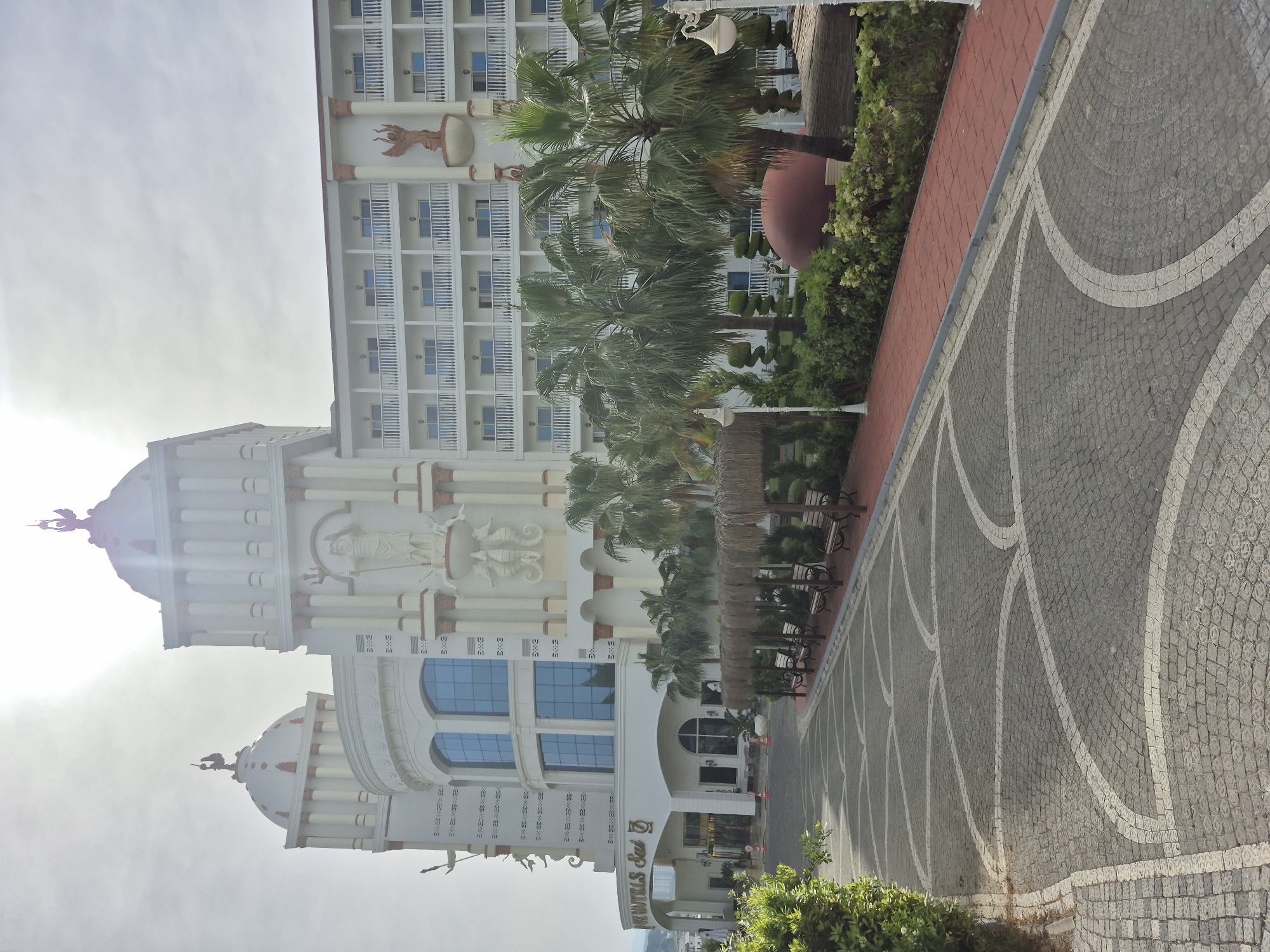 The image depicts the exterior of the Lonicera Resort & Spa hotel in Alanya, Turkey. The hotel has a white facade with neoclassical architectural elements, including columns, domes, and statues of mythological figures. Palm trees are planted in front of the hotel. A paved walkway with a decorative pattern runs along the side of the building. Benches are placed along the walkway. The sky is overcast.