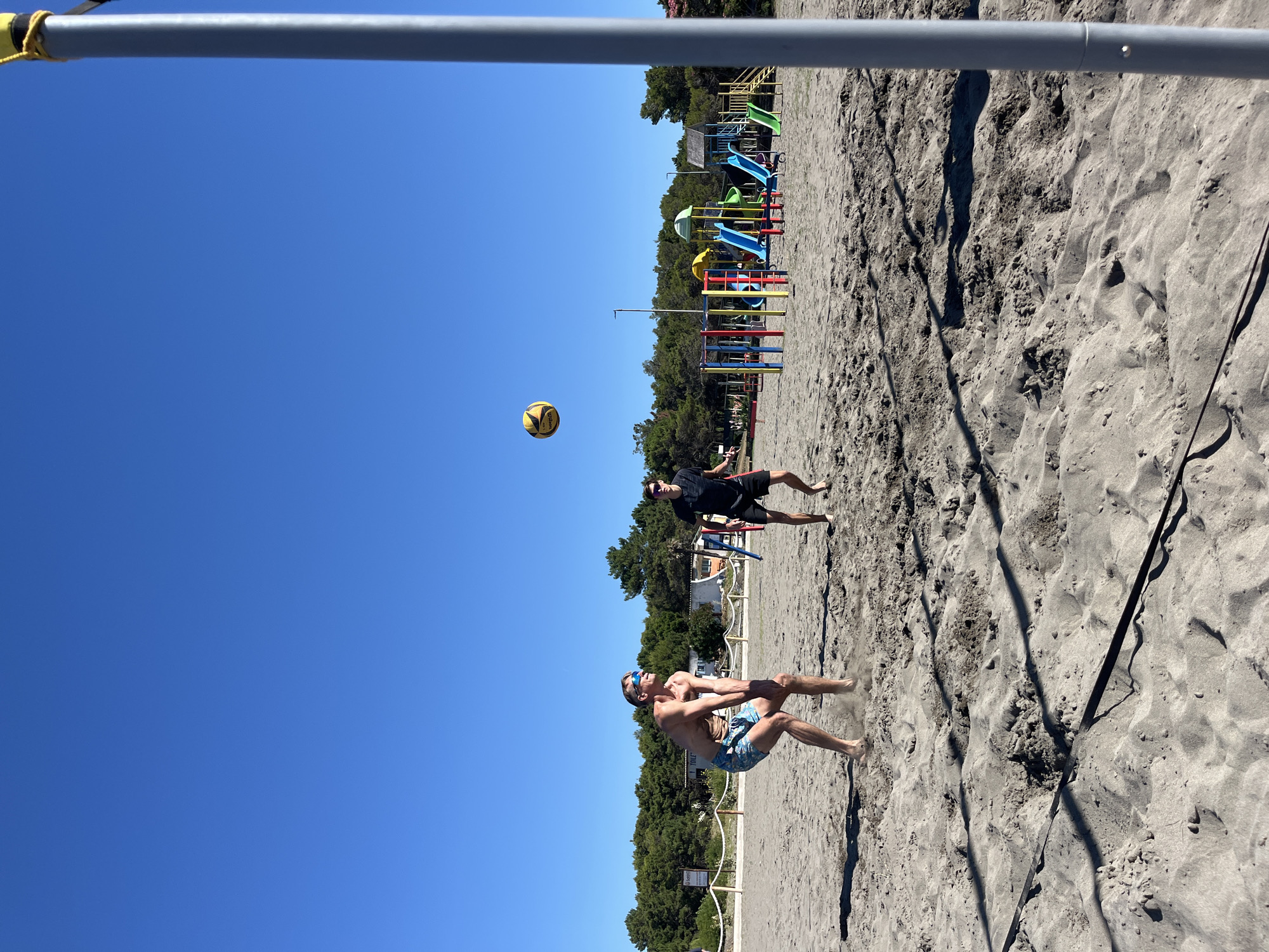 Two men are playing volleyball on a sandy beach. The man in the foreground is shirtless and wearing patterned swim trunks, in a crouched position ready to receive the ball. The man in the background is wearing a black t-shirt and black shorts, and is looking up at the ball. The volleyball is in mid-air, yellow and black. In the background, there is a playground with slides and climbing structures. A line of trees separates the beach from buildings. The sky is a clear, bright blue. The image appears to be taken on a sunny day. The location is the beach in Pistula, Montenegro, known for its sandy shores and proximity to playgrounds.