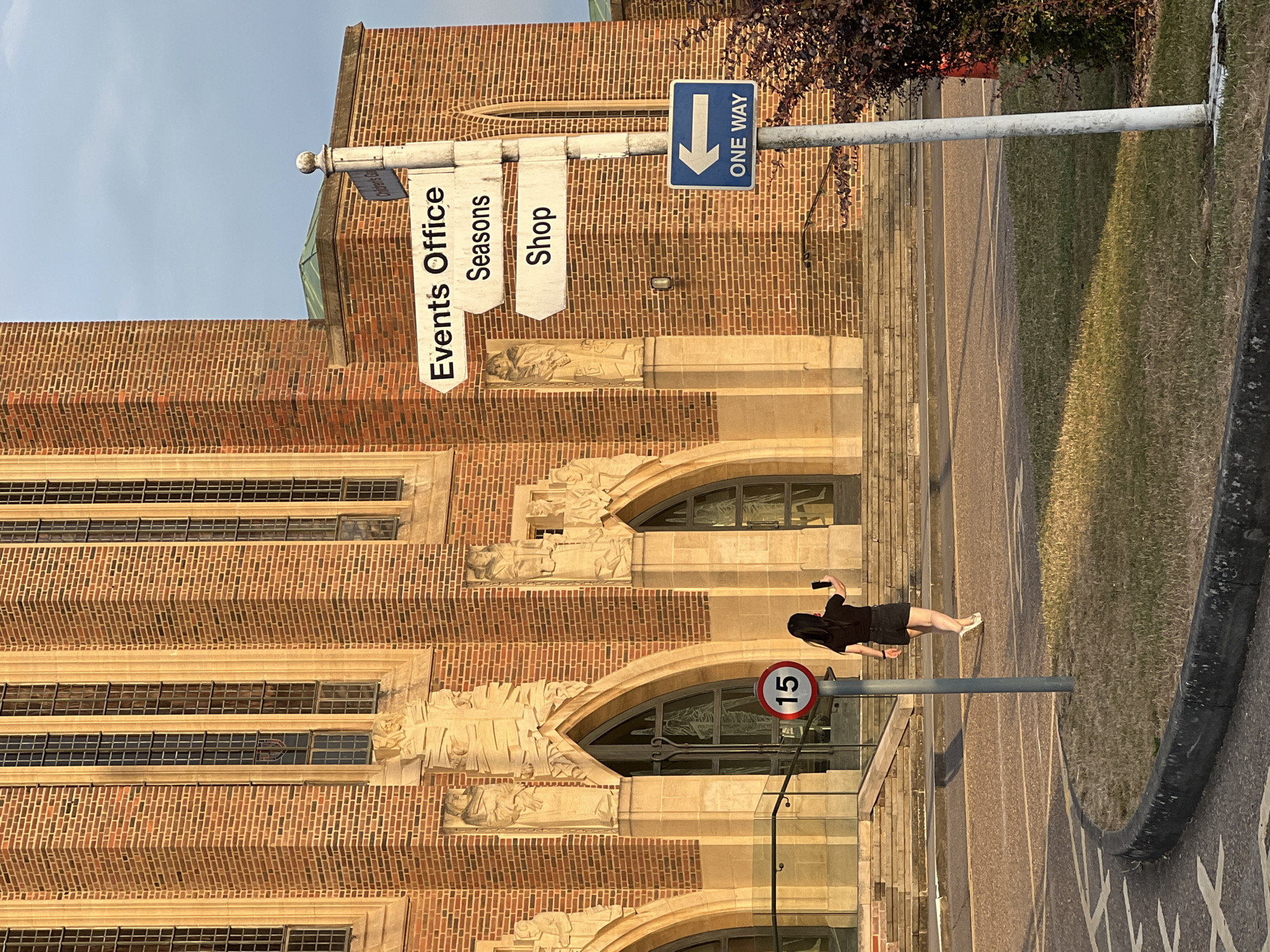 A woman stands outside a brick building with large arched windows. She is dressed in a black top and skirt and is holding a phone in her right hand. The building appears to be a public building, possibly a civic center or university. There are signs for "Events Office", "Seasons", and "Shop" hanging from a pole. A "ONE WAY" sign with a blue arrow pointing down is also visible. A speed limit sign showing "15" is on another pole. The setting appears to be outdoors, possibly in the late afternoon given the lighting and shadows. The sky is clear and blue. The building's architecture features intricate carvings above the arched windows. The grass area next to the building suggests it is a public space. The image is oriented vertically.