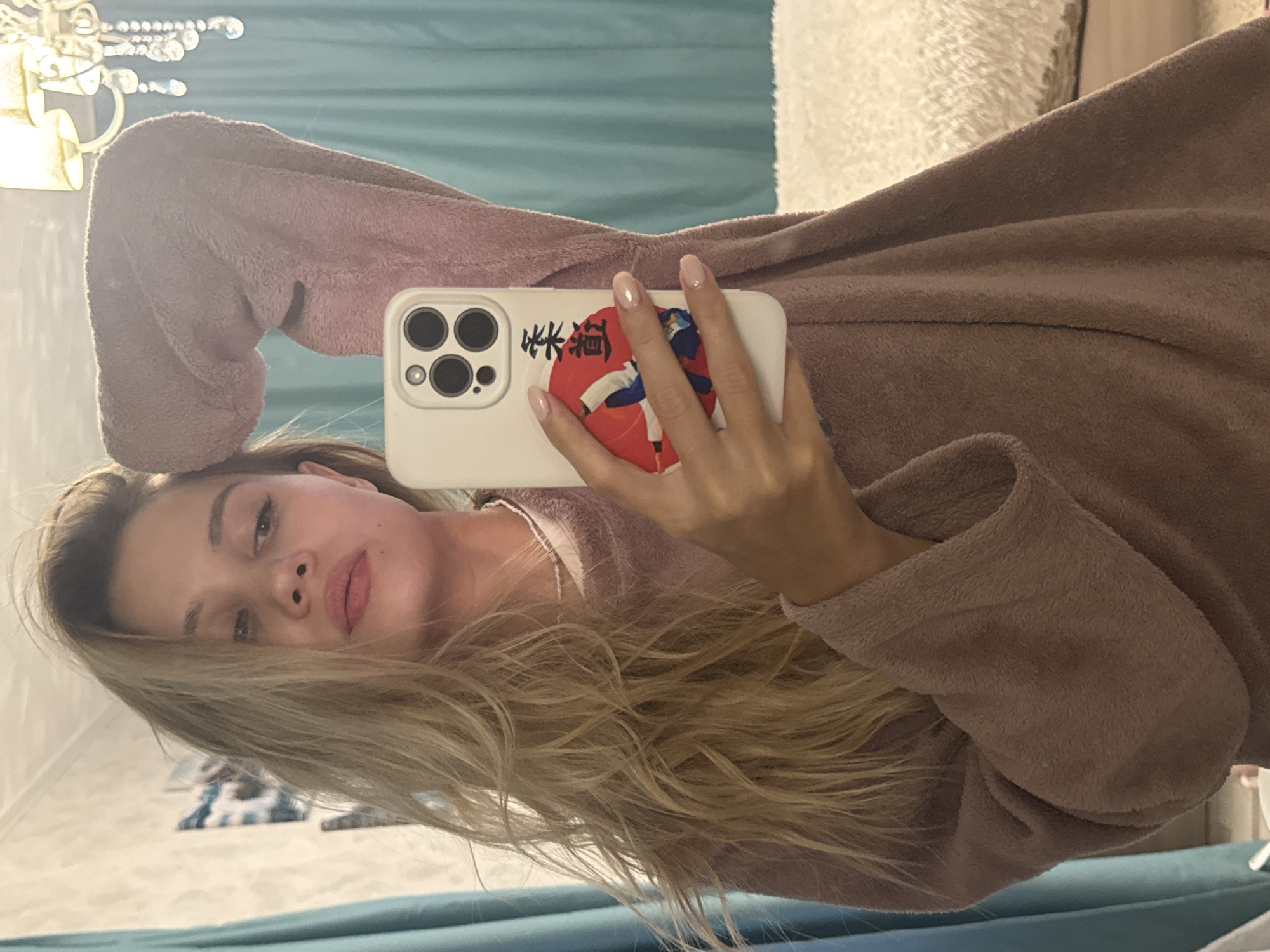 A person with blonde hair and light skin holds a white smartphone with a red and white sticker on the back, likely taking a selfie. They are wearing a mauve robe or dressing gown. The background includes a turquoise curtain and a glimpse of a white patterned surface. The location is Chaikovsky, Russia.