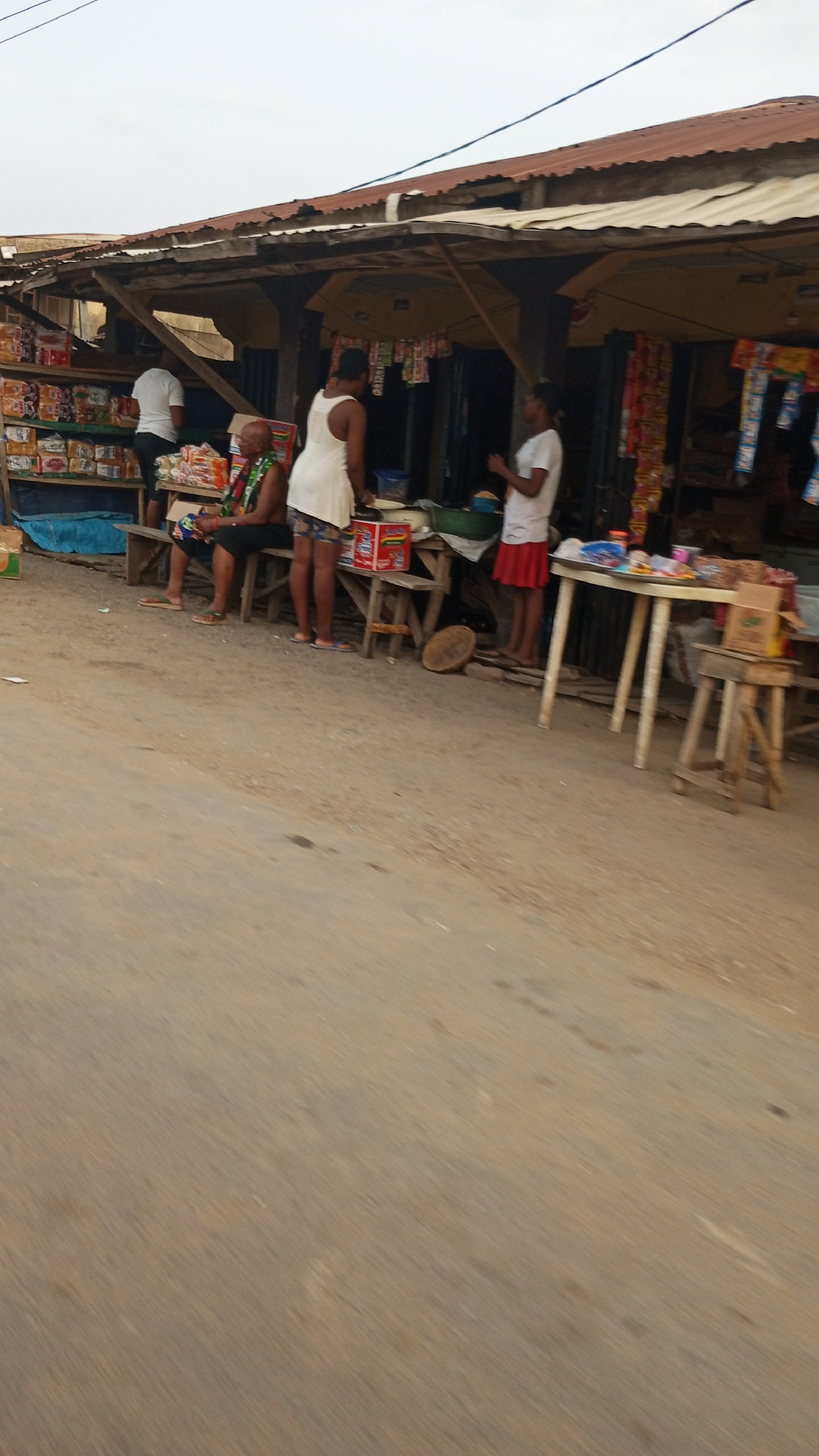 The image depicts a street scene in Adankolo, Nigeria. Several individuals are present in front of a roadside shop. A person in a white shirt stands within the shop. Two women are standing in front of the shop, one wearing a white dress and the other a white shirt and red skirt. An older individual sits on a bench to the left of the shop. Various goods are displayed for sale within the shop, including packaged foods and snacks. Tables and stools are positioned in front of the shop, holding additional items for sale. The structure of the shop appears rudimentary, with a corrugated metal roof. The road is unpaved.