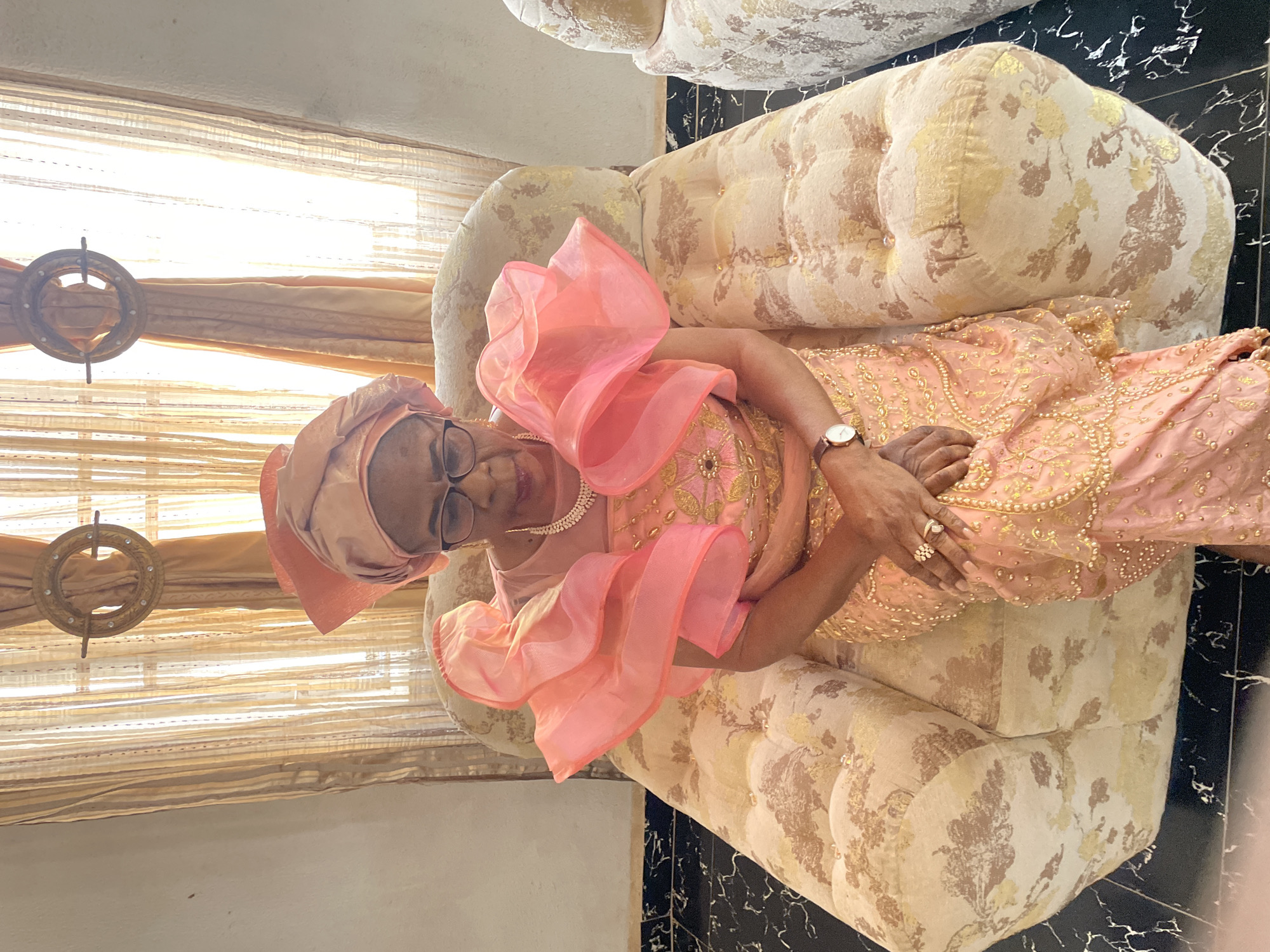 An elderly woman is sitting on an ornately patterned upholstered sofa, indoors. She is wearing an elaborate peach-colored outfit with ruffles at the shoulders and a matching headwrap. She has a pearl necklace and a watch on her wrist. Her hands are folded in her lap, and rings are visible on her fingers. Behind her, a window is covered with tan-colored, semi-sheer blinds and a decorative wooden ring. The floor has a black and white marble pattern. Location: Unknown, Unknown.