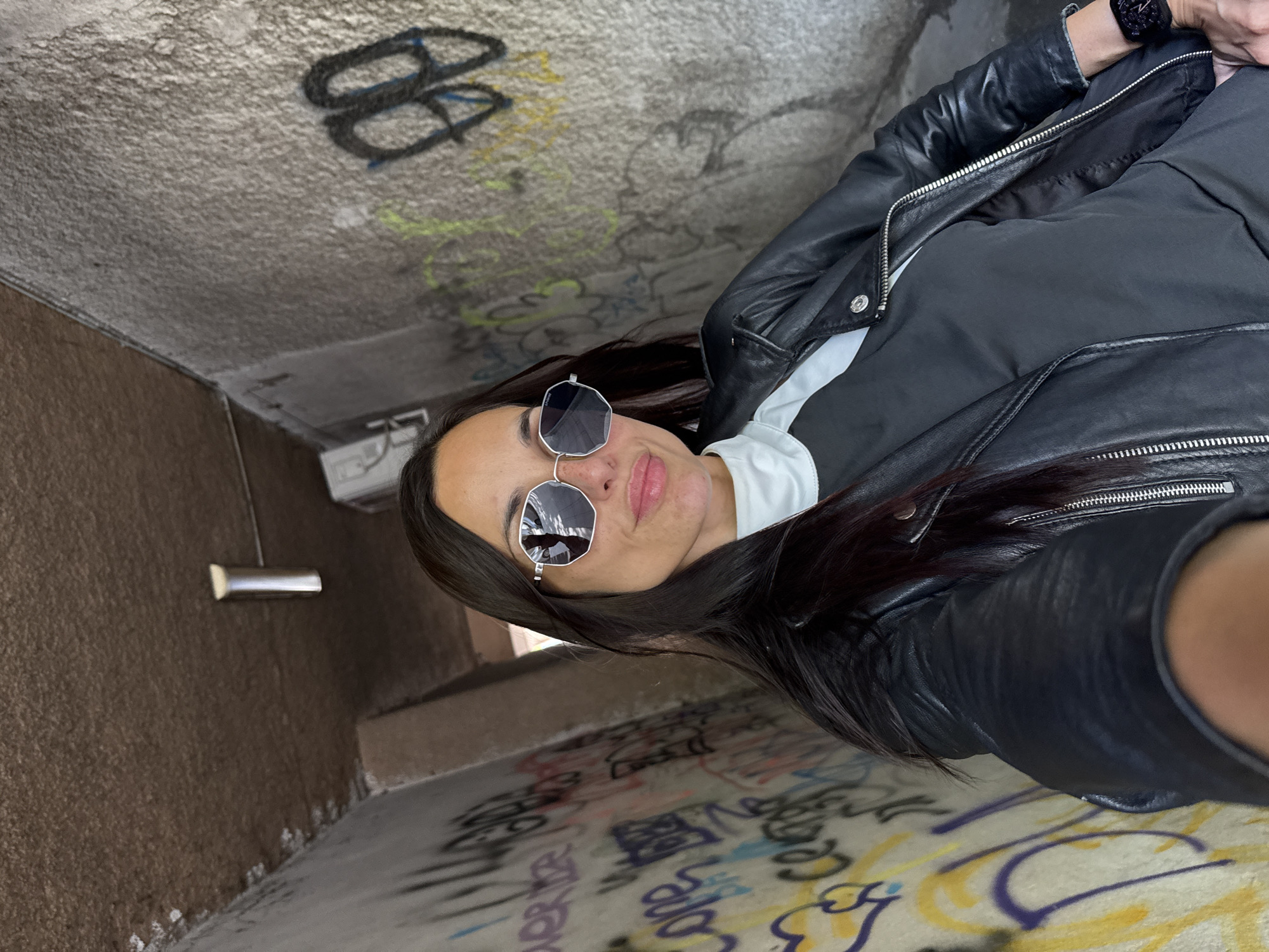 A woman is taking a selfie in what appears to be a graffiti-covered underpass or tunnel in Pleven, Bulgaria. The graffiti is extensive and covers the walls and ceiling. The woman is wearing sunglasses and a black leather jacket over a white collared shirt and a dark t-shirt. Her hair is long and dark. She is wearing a wristwatch. The light is dim, and the environment seems urban and potentially gritty.