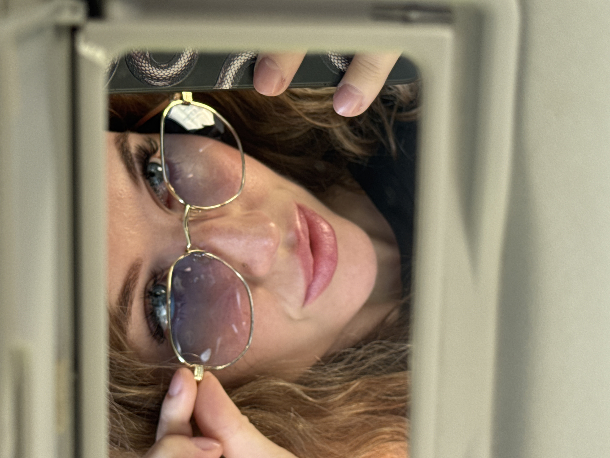 A woman is seen taking a selfie in a car's visor mirror. She is wearing gold-rimmed sunglasses with gradient lenses and has light brown, curly hair. Her lips are painted a soft pink. She is holding the sunglasses with her left hand and a smartphone with a snake pattern on its case with her right hand. The background is out of focus, but appears to be the interior of a car. The image was taken in Chaikovsky, Russia.
