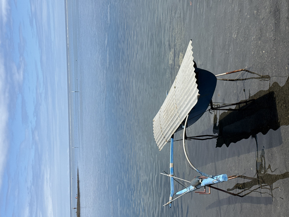 The scene depicts a calm, shallow coastal area in Māhina, France, under a clear sky with scattered white clouds. In the foreground, partially submerged in clear, blue-green water, are two improvised structures. The structure on the left consists of a blue, log-like beam supported by rusty metal poles forming a tripod, with a smaller blue cross-beam and a dark cylindrical object attached. The structure on the right features a dark blue, inverted trough or hull-like object covered by a corrugated galvanized metal sheet, secured by rope, and also supported by rusty metal poles. Both structures cast dark, elongated shadows onto the visible sandy seabed. In the midground, the calm water extends, revealing two slender white poles protruding vertically. Further in the background, a low-lying landmass or sandbar is visible on the left, with three indistinct human figures standing on it. Beyond this, a faint white line indicates a distant reef or wave break, separating the calm water from the open sea under a hazy horizon.